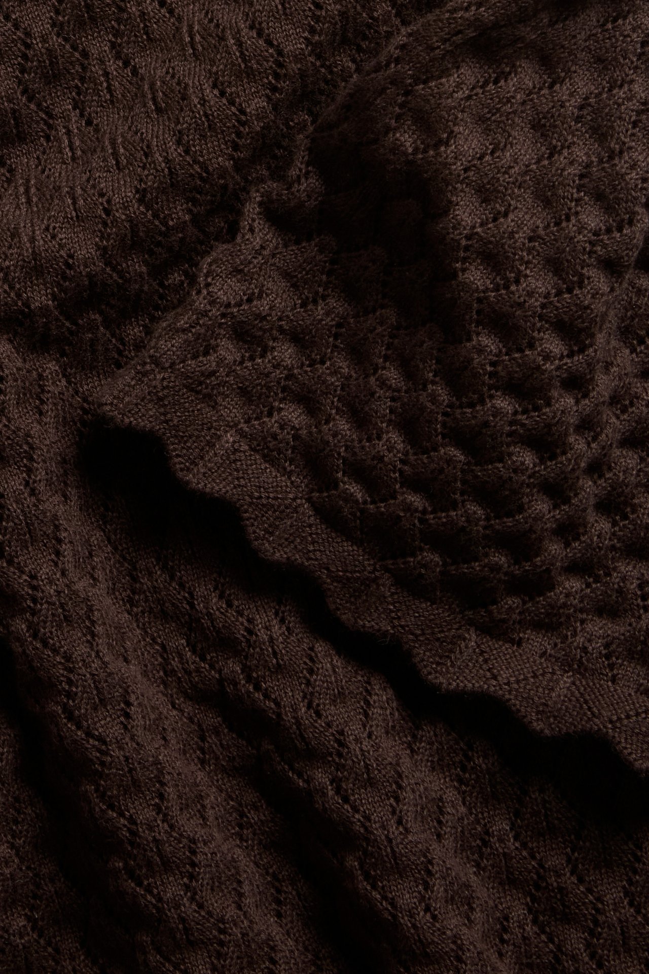 Närbild av en mörkbrun mönsterstickad kortärmad bomullströja med hålmönster och vågig kant.