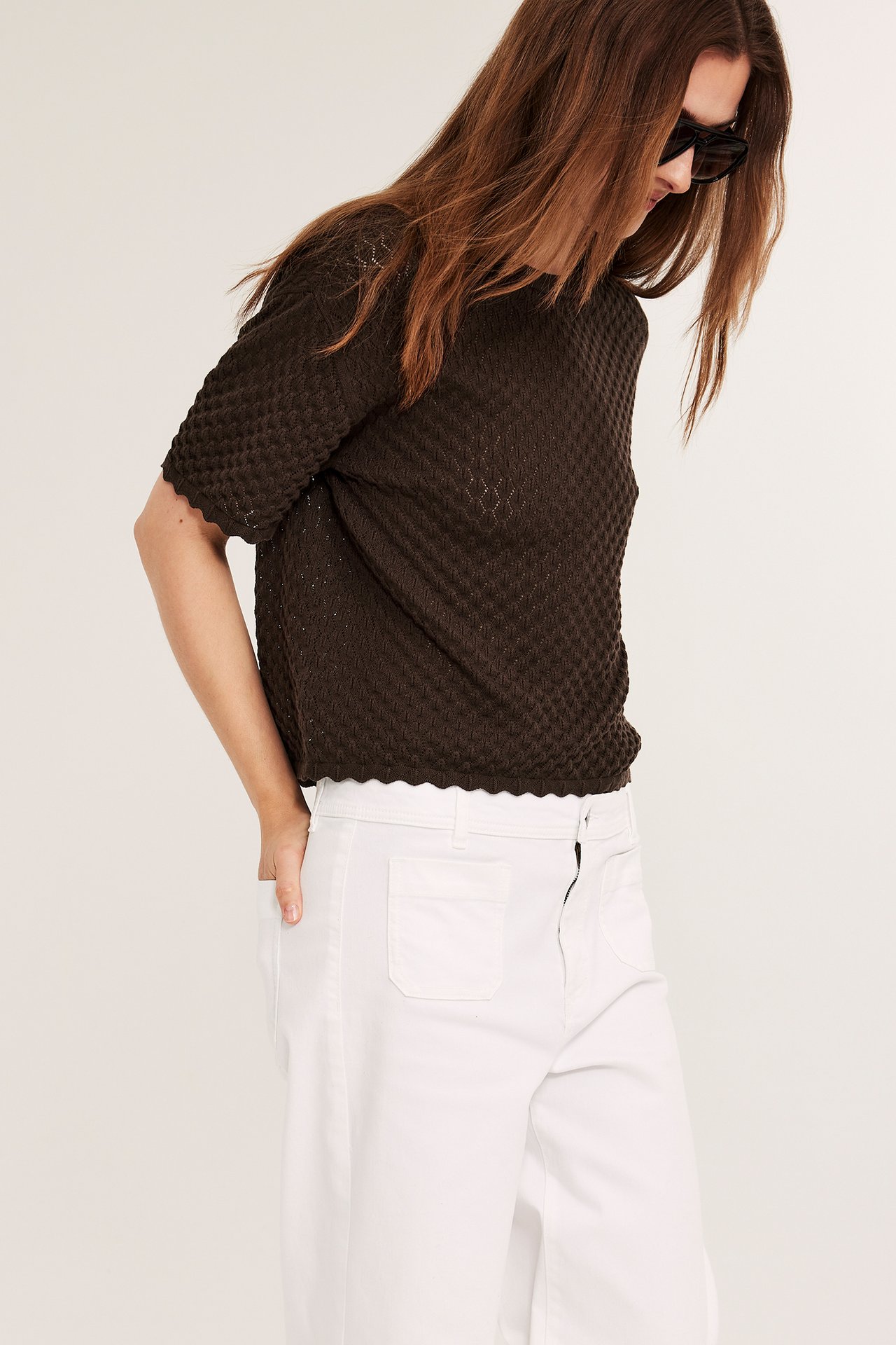 Edestä: nainen yllään tummanruskea lyhythihainen neuletoppi, valkoiset leveälahkeiset housut ja mustat aurinkolasit.