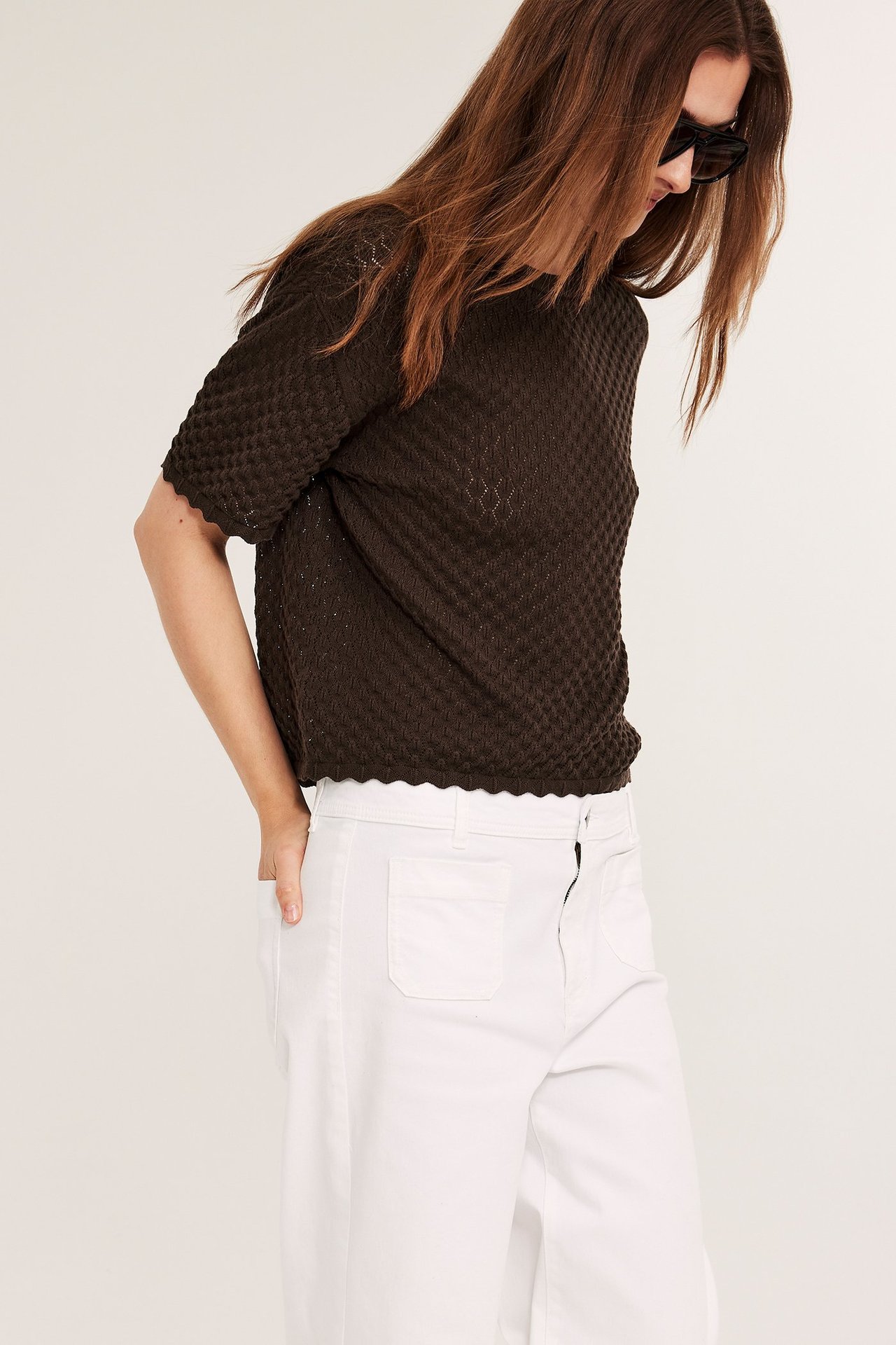 Widok z przodu: kobieta w ciemnobrązowej dzianinowej bluzce z krótkim rękawem, białych spodniach i czarnych okularach.