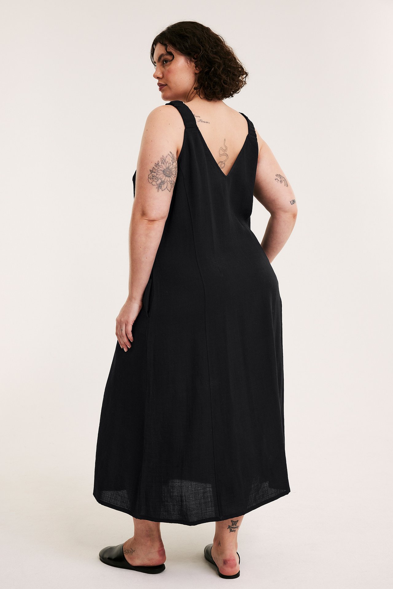 Bakre sidovy av kvinna i lång svart viskosklänning med V-rygg och sidofickor, bär svarta skor.