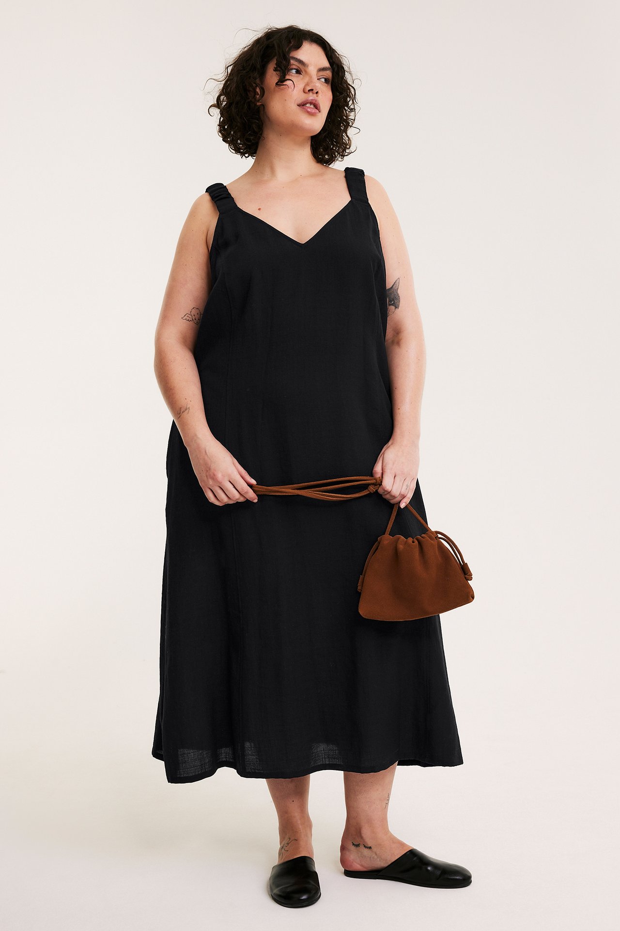 Framifrån: kvinna i lång svart V-ringad viskosklänning, håller en brun dragskoväska och svarta slip-on skor.