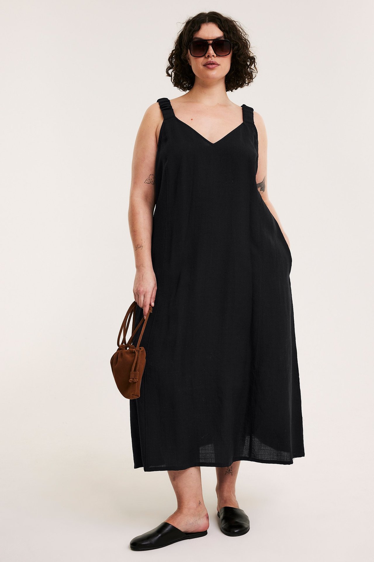 Frontbild: Kvinna i svart lång viskosmixklänning med V-ringning, solglasögon, håller brun väska, bär svarta mules.