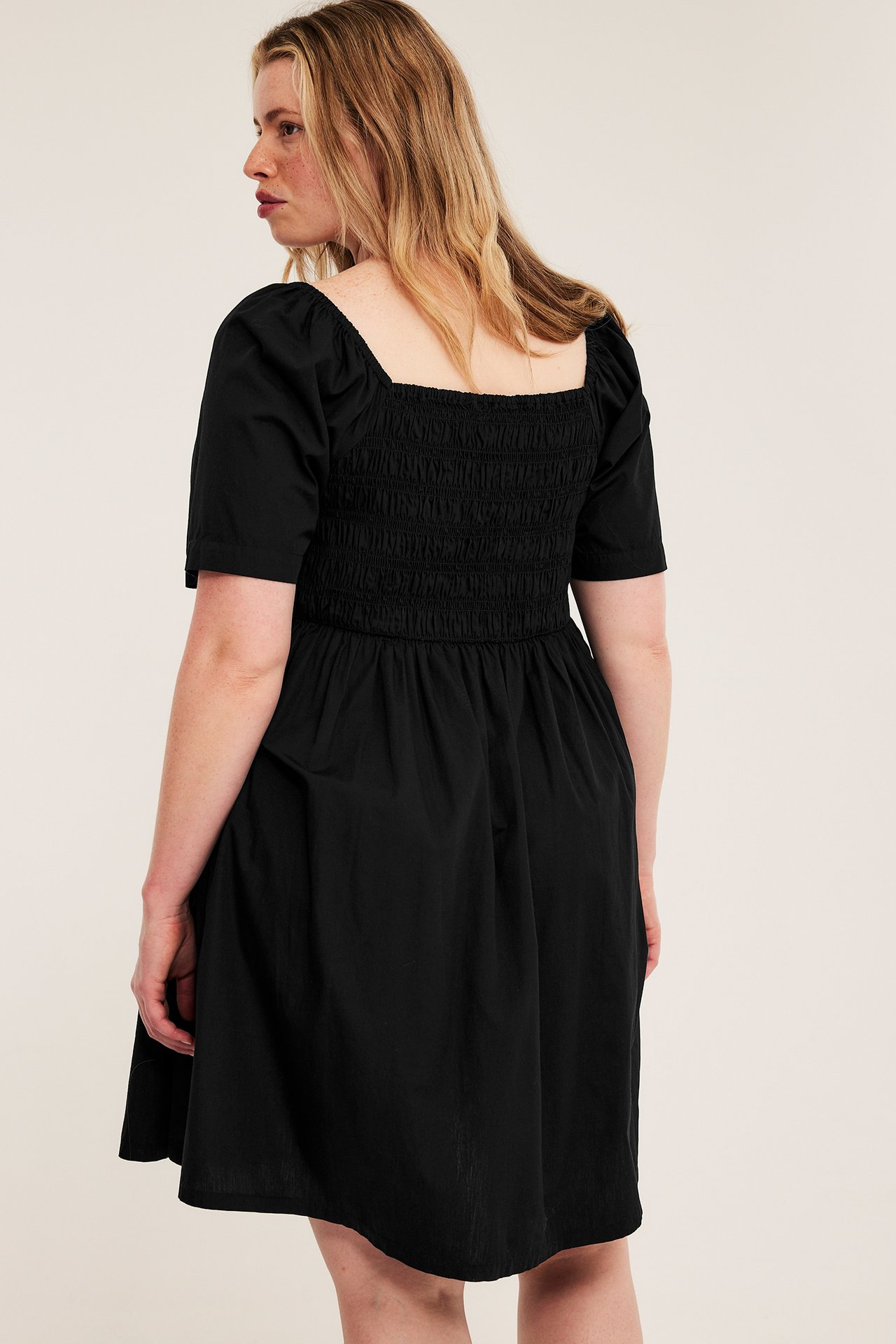 Baksidan av en kvinna i svart poplinklänning med smockat livstycke, fyrkantig ringning och puffärmar.