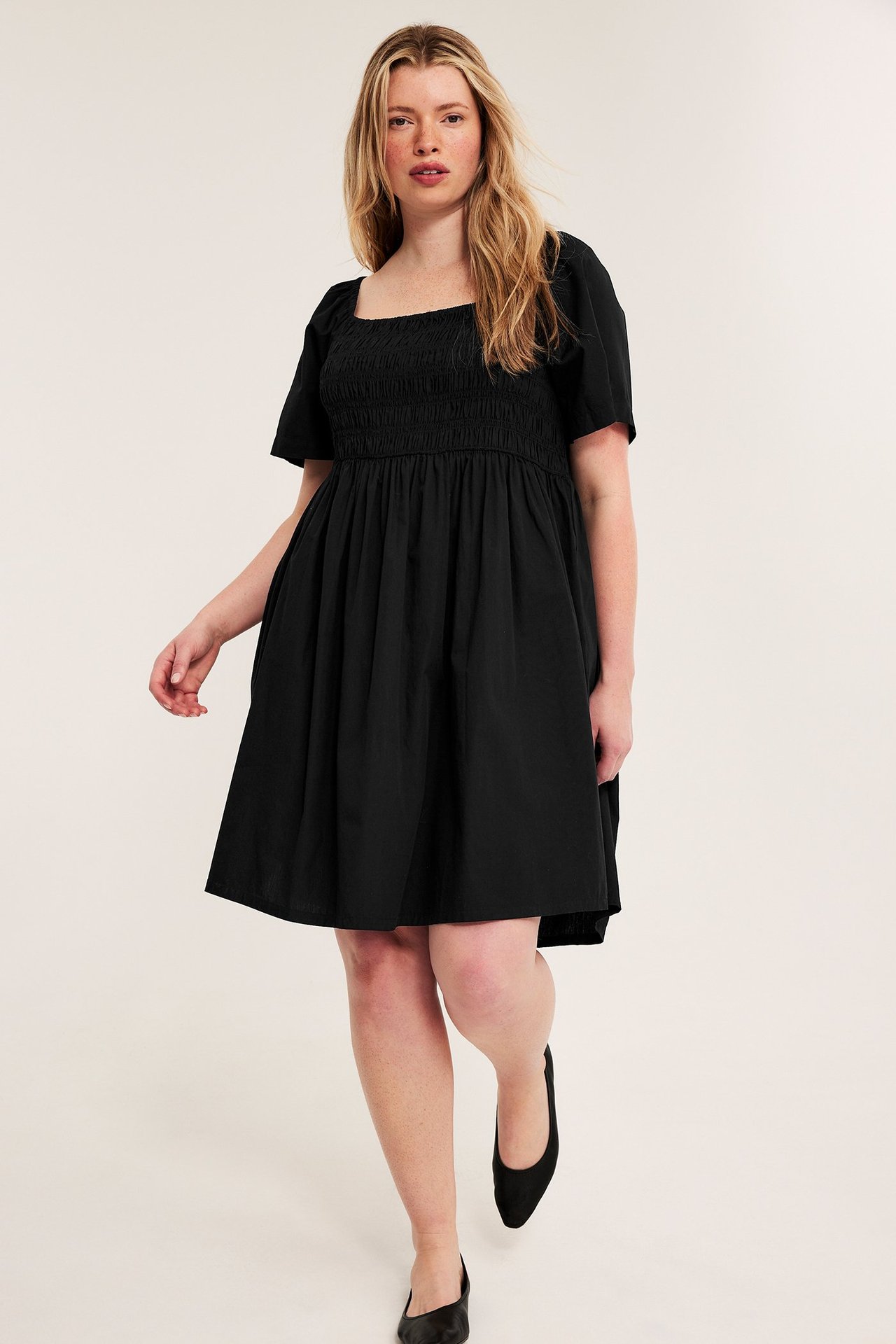 Framifrån: kvinna i svart poplinklänning med smock, puffärmar och svarta platta skor.