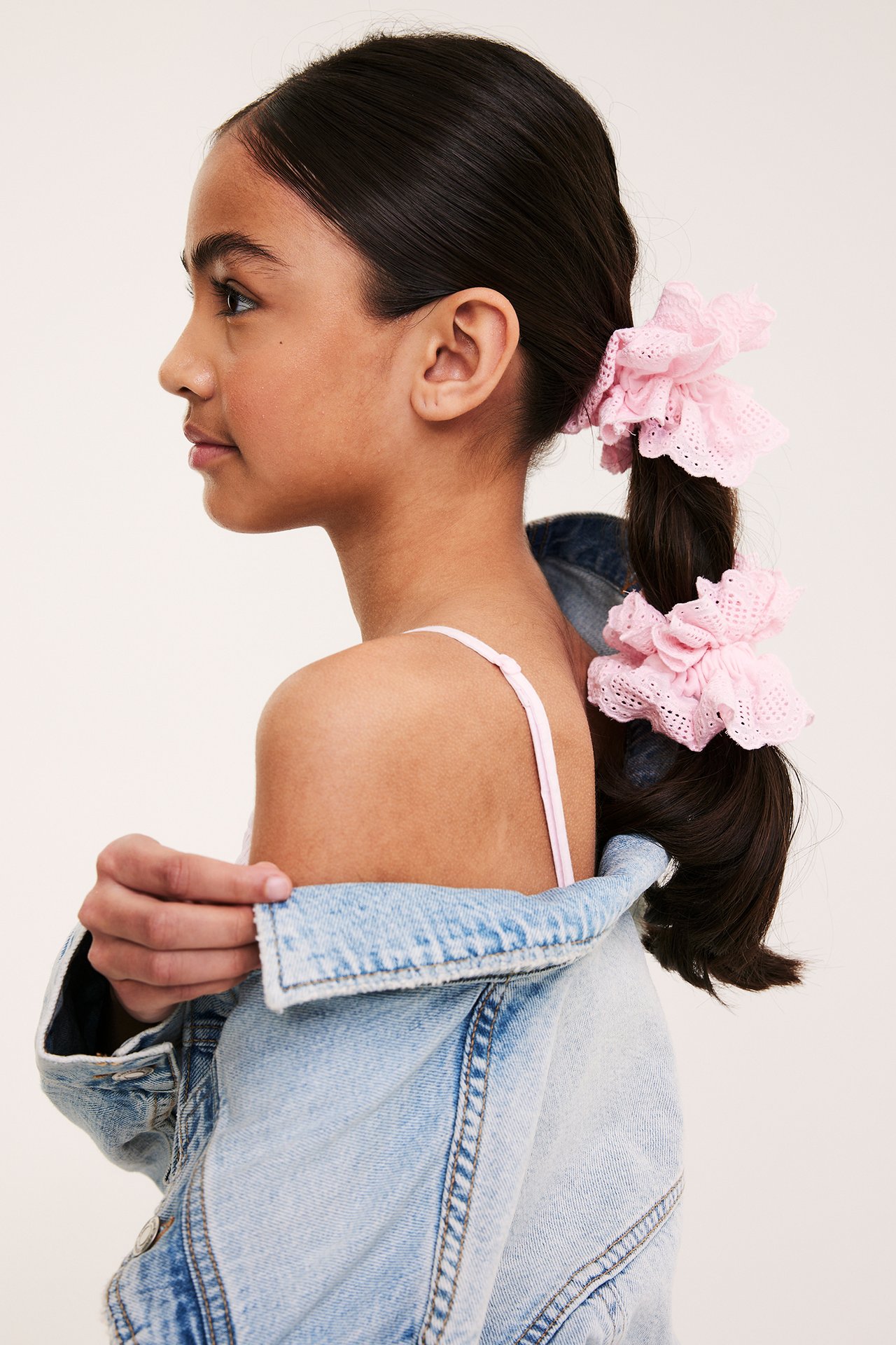 Widok z boku dziecka z dwiema różowymi frotkami z haftem angielskim w kucyku, noszącego jasnoniebieską kurtkę jeansową.