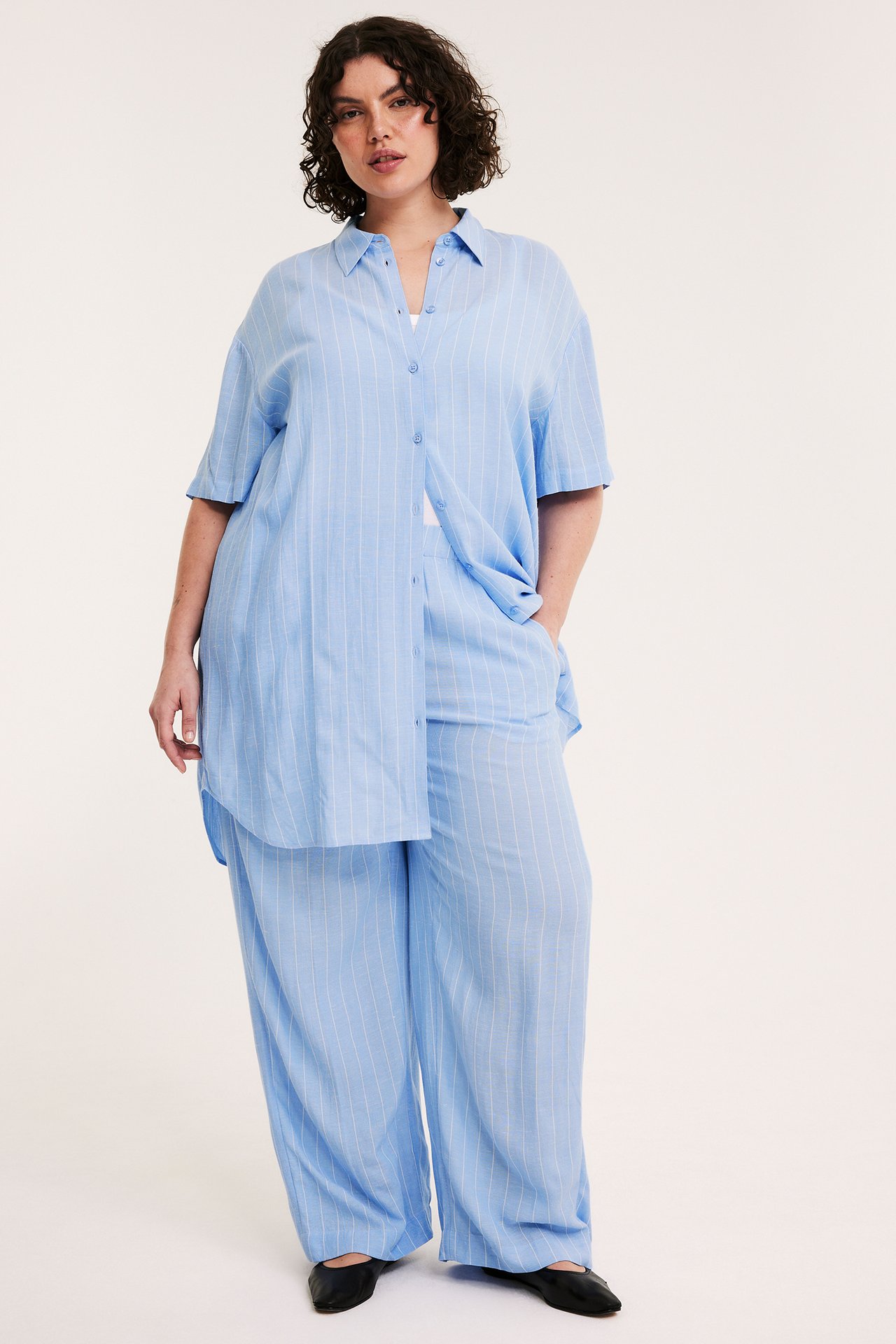 Front view: Kvinna i ljusblå randig långskjorta i viskos, matchande byxor och svarta platta skor.
