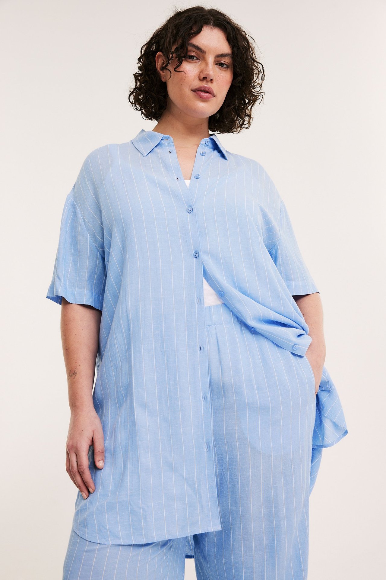 Front: Kvinna i öppen ljusblå randig långskjorta över vit topp, med matchande byxor.