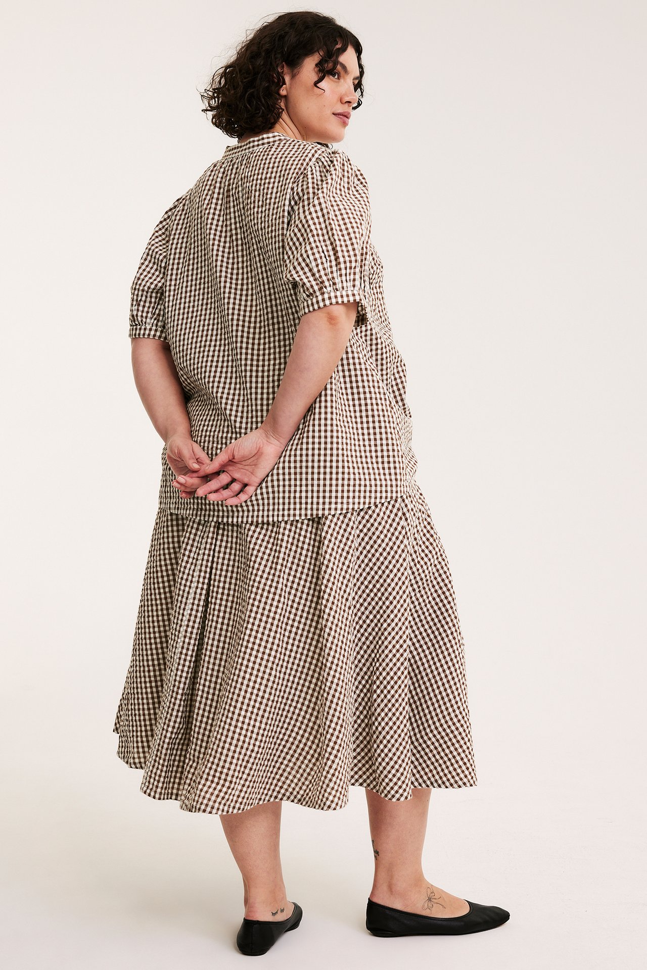 Selkä: nainen ruskearuudullisessa seersucker-puserossa, puffihihat, yhteensopiva hame, mustat ballerinat.
