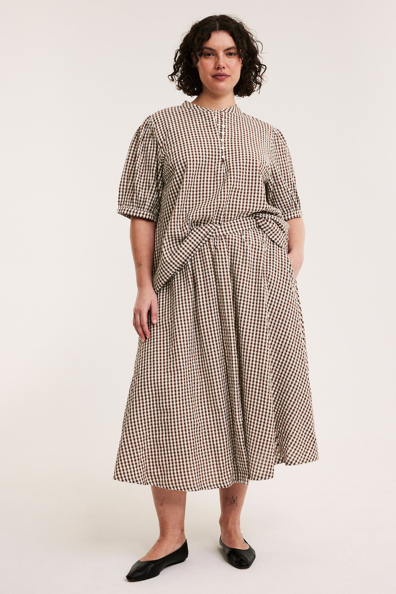 Nainen ruskeassa ruudullisessa seersucker-puserossa, pussihihoilla ja yhteensopivassa hameessa. Mustat kengät.