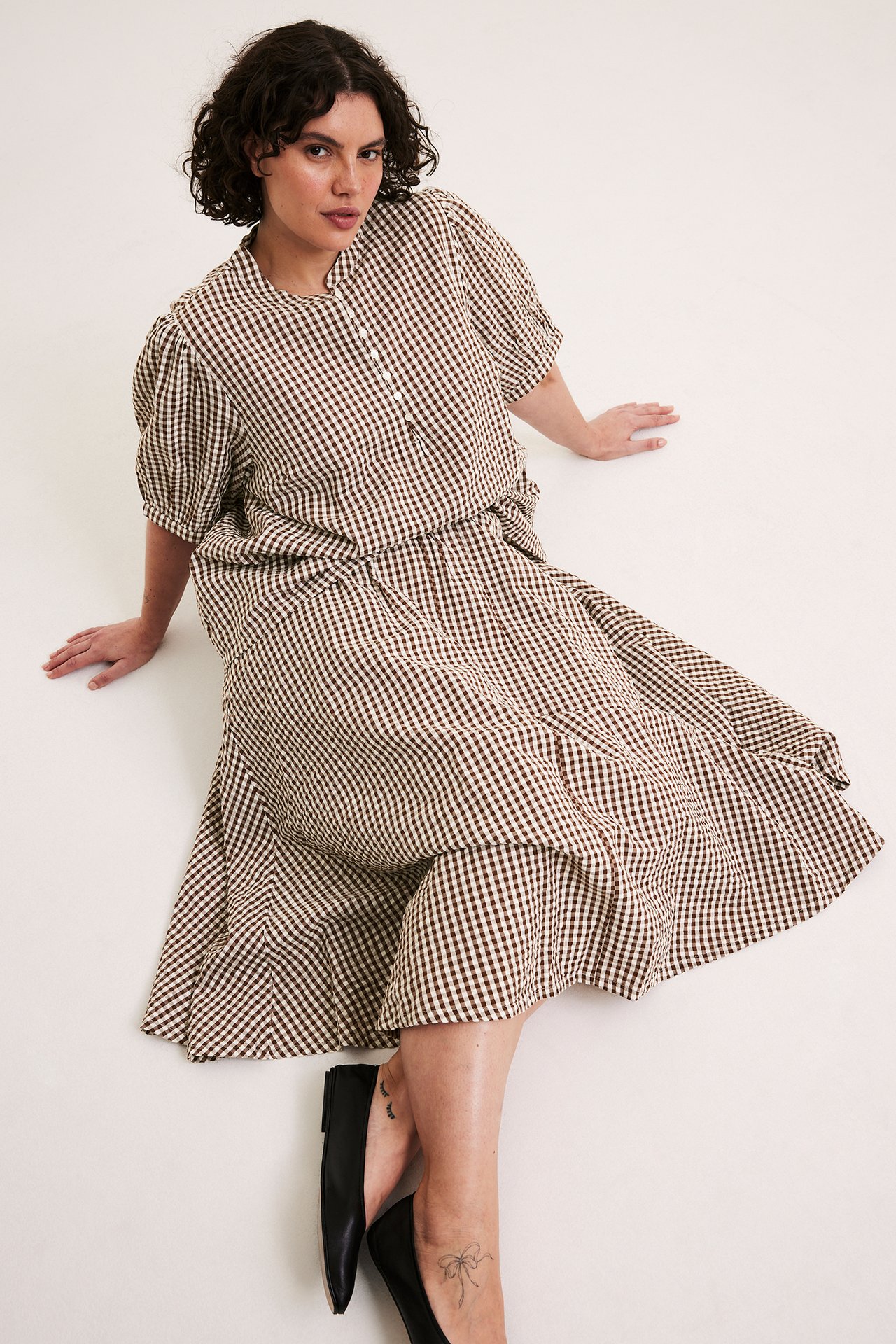Edestä: nainen ruskeassa ruudullisessa puuvillapaidassa, jossa pussihihat, yhteensopiva hame ja mustat kengät.