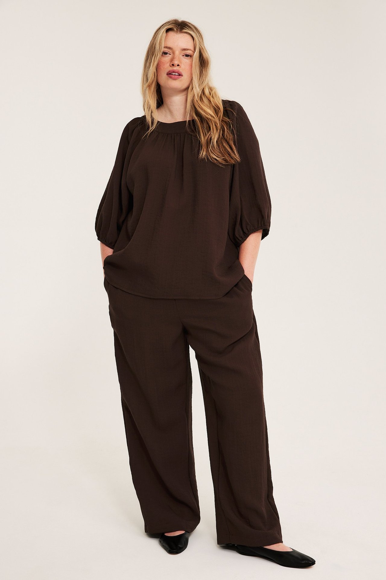 Edestä: Nainen pukeutuu tummanruskeaan Singoalla-puseroon, jossa puffihihat, vastaavat leveälahkeiset housut ja mustat ballerinat.