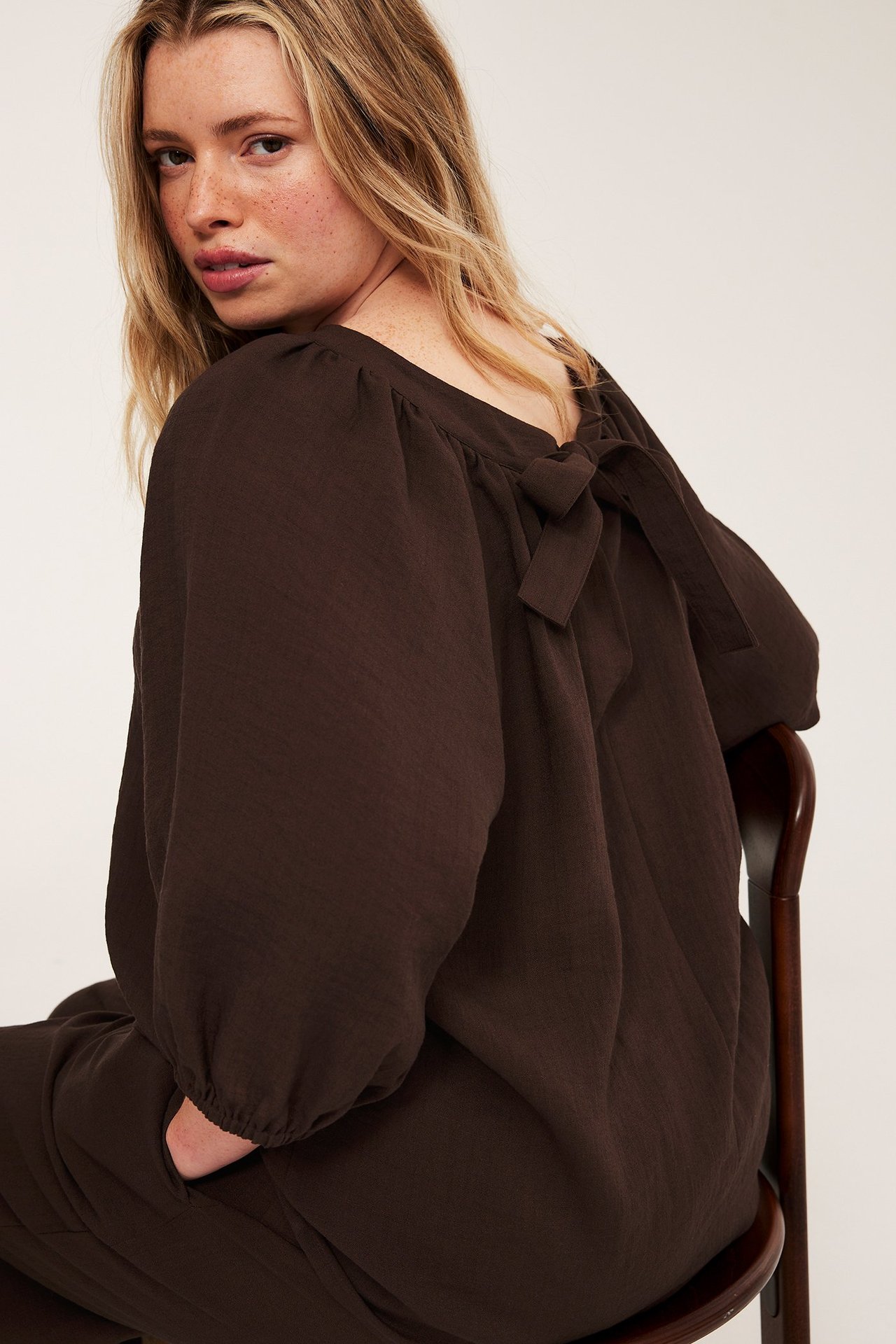 Nainen tummanruskeassa Singoalla-puserossa, jossa pitkät pussihihat ja rusetti takana, yllään vastaavat housut.