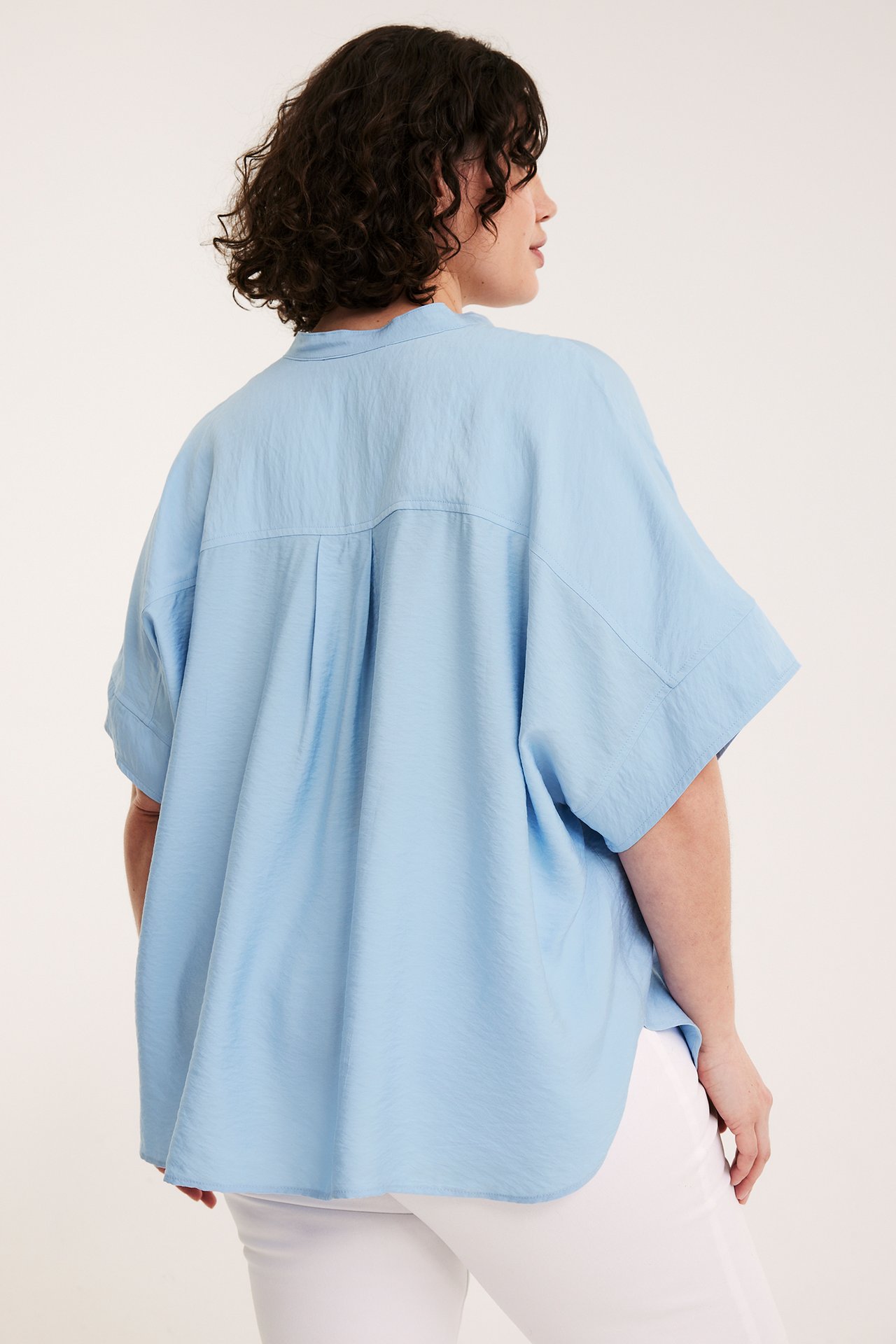 Widok z tyłu/boku kobiety w jasnoniebieskiej, luźnej bluzce popover z krótkim rękawem i białych spodniach.