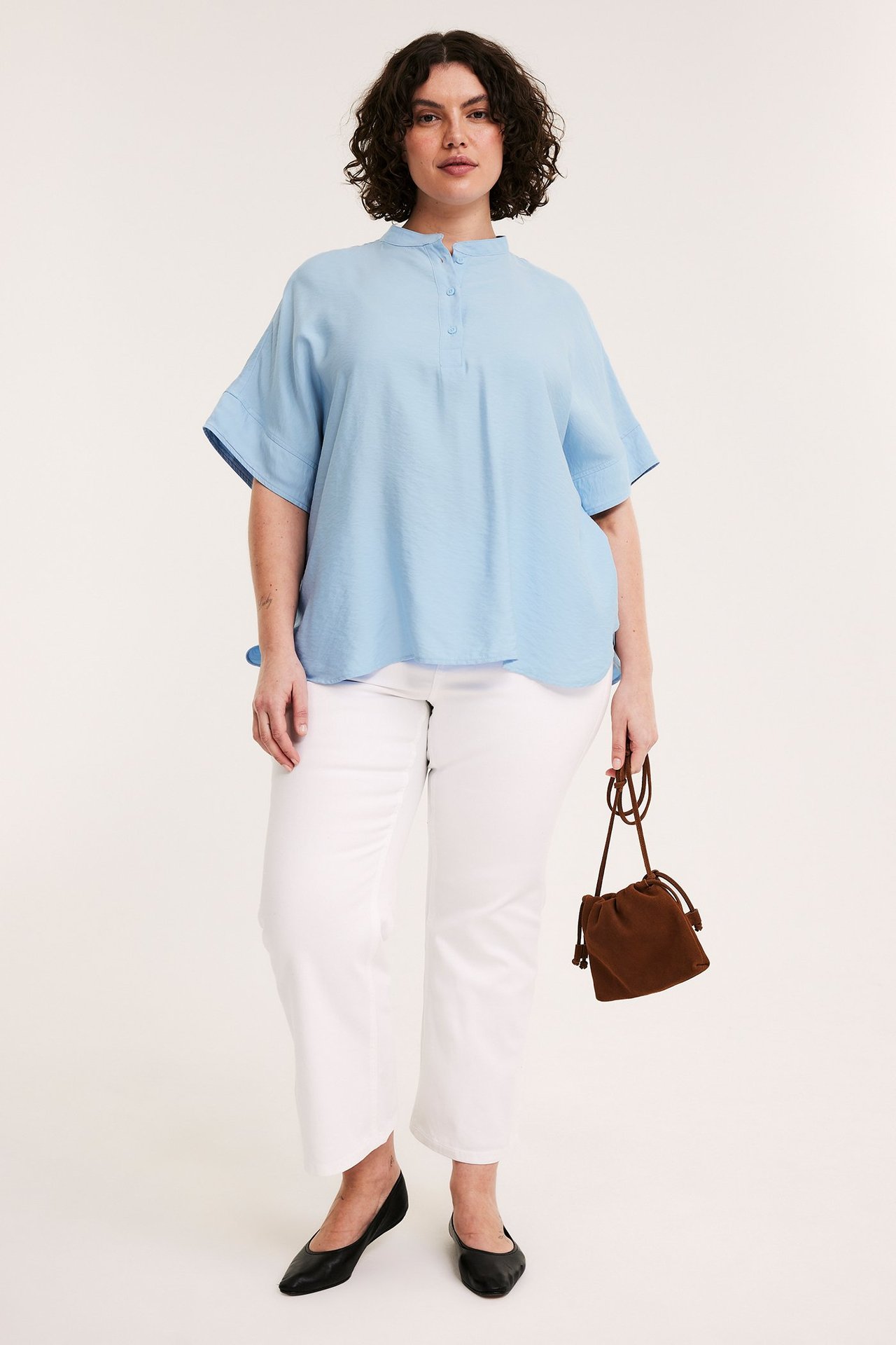 Z przodu: kobieta w jasnoniebieskiej bluzce popover, białych spodniach, czarnych baletkach, z brązową torebką.