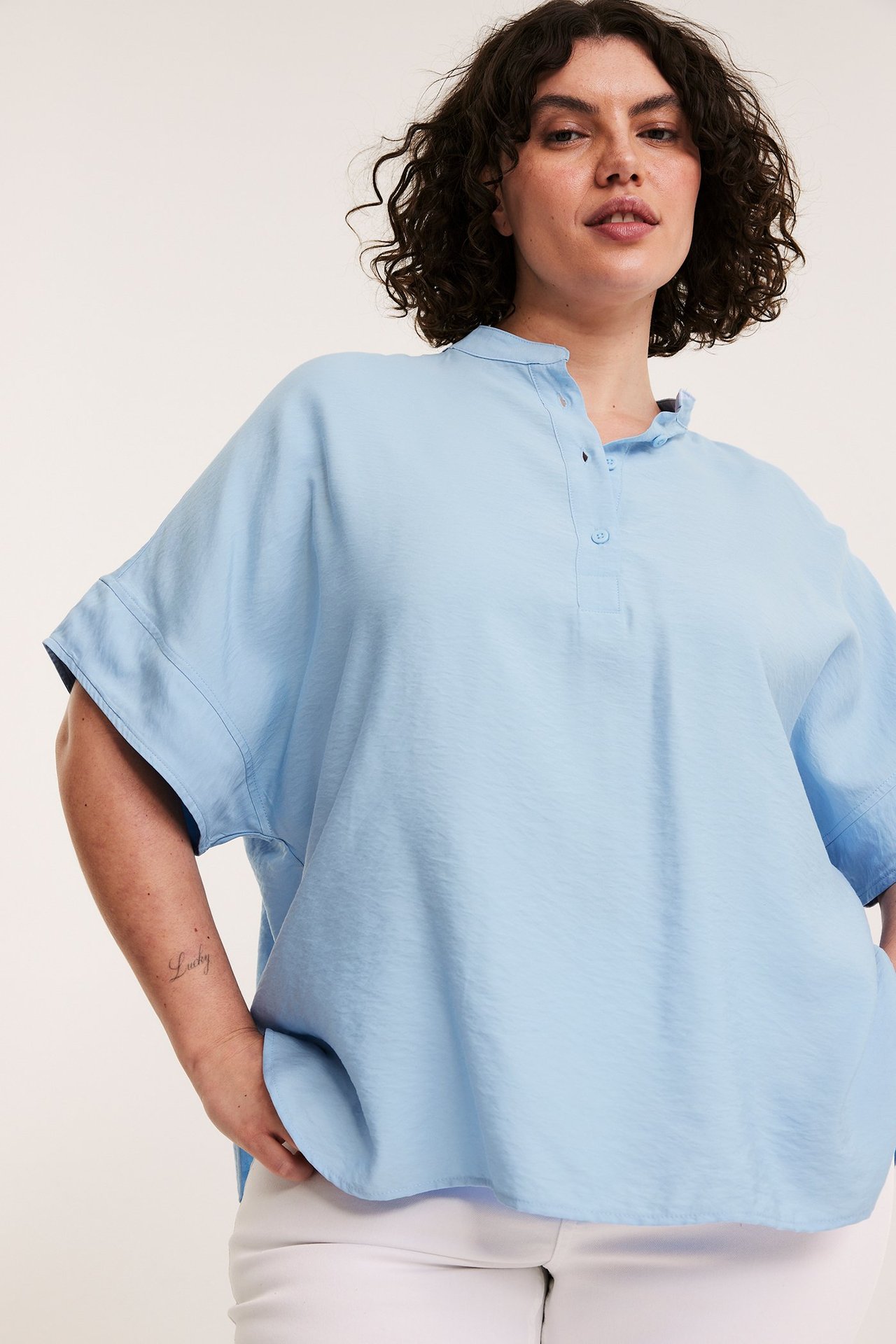 Widok z przodu: Kobieta w błękitnej, obszernej bluzce popover bez kołnierza z krótkimi rękawami i białych spodniach.