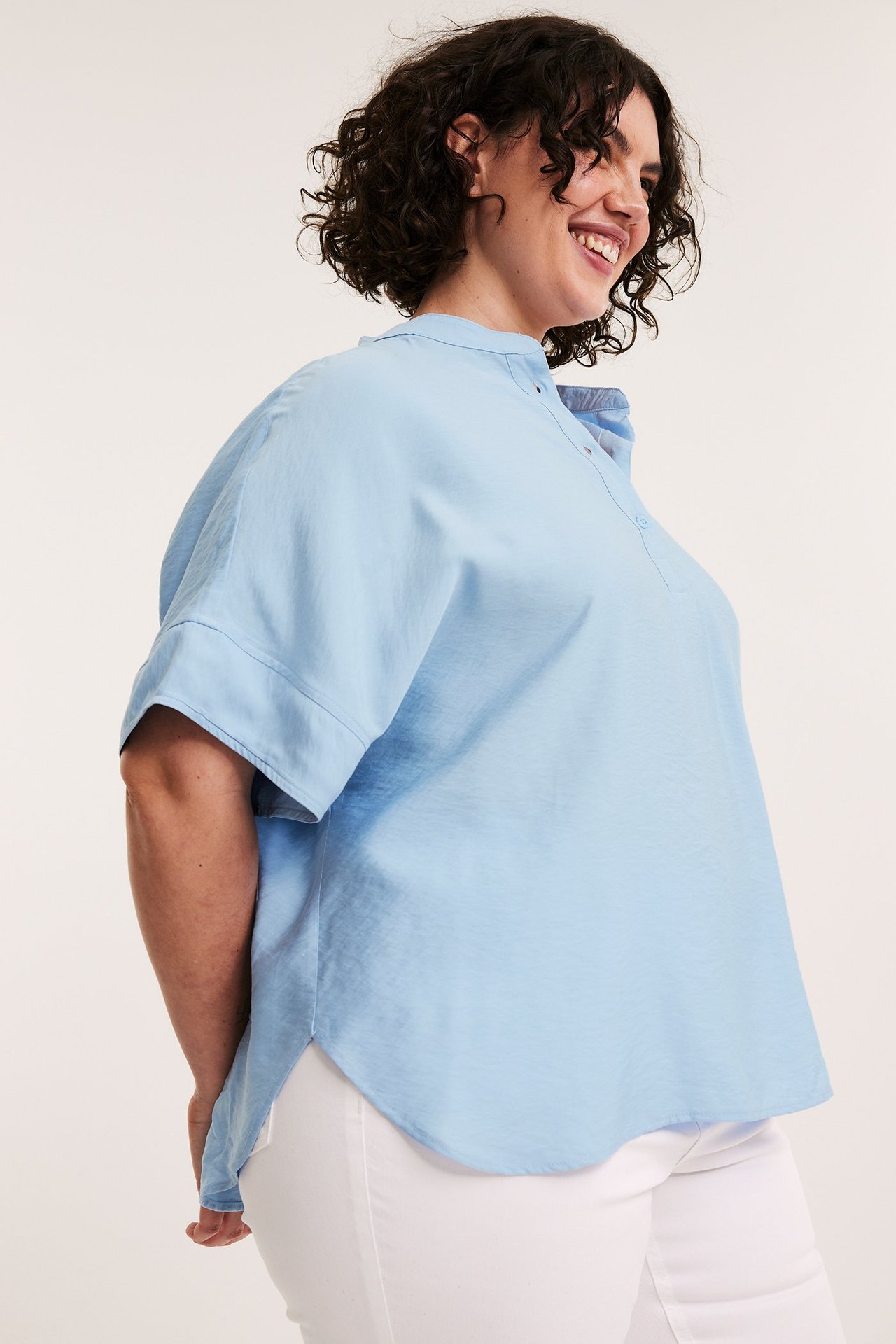 Kvinne i lys blå kortermet popover-bluse med avrundet kant og hvite bukser, sidevisning.