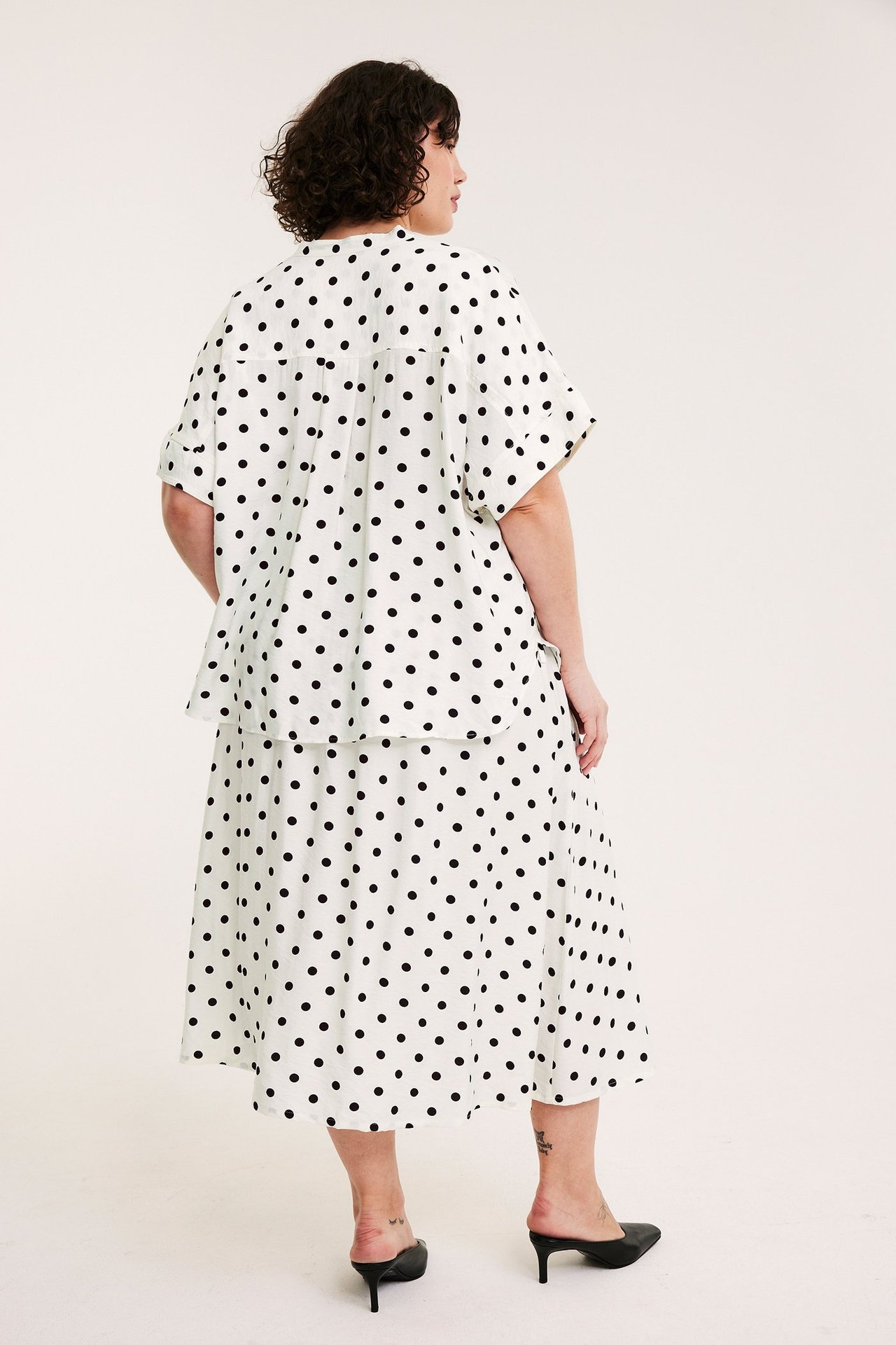 Widok z tyłu kobiety w luźnej bluzce popover w kropki, dopasowanej spódnicy i czarnych klapkach.