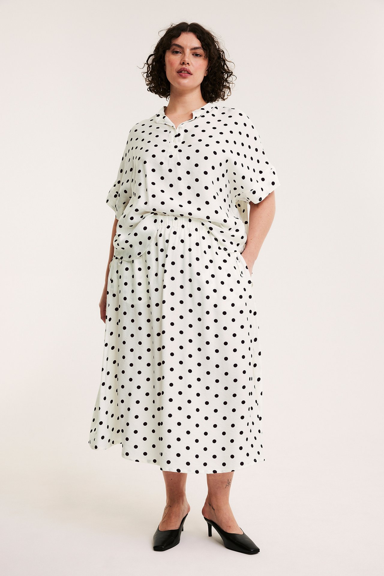 Forfra: Kvinne i kremhvit prikkete popoverbluse med droppet erme, matchende skjørt og svarte mules.