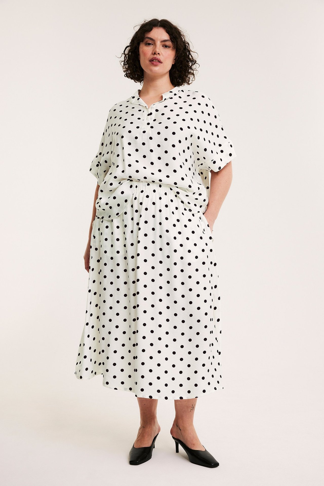Edestä: Nainen luonnonvalkoisessa pilkullisessa popover-puserossa, yhteensopivassa hameessa ja mustissa muuleissa.