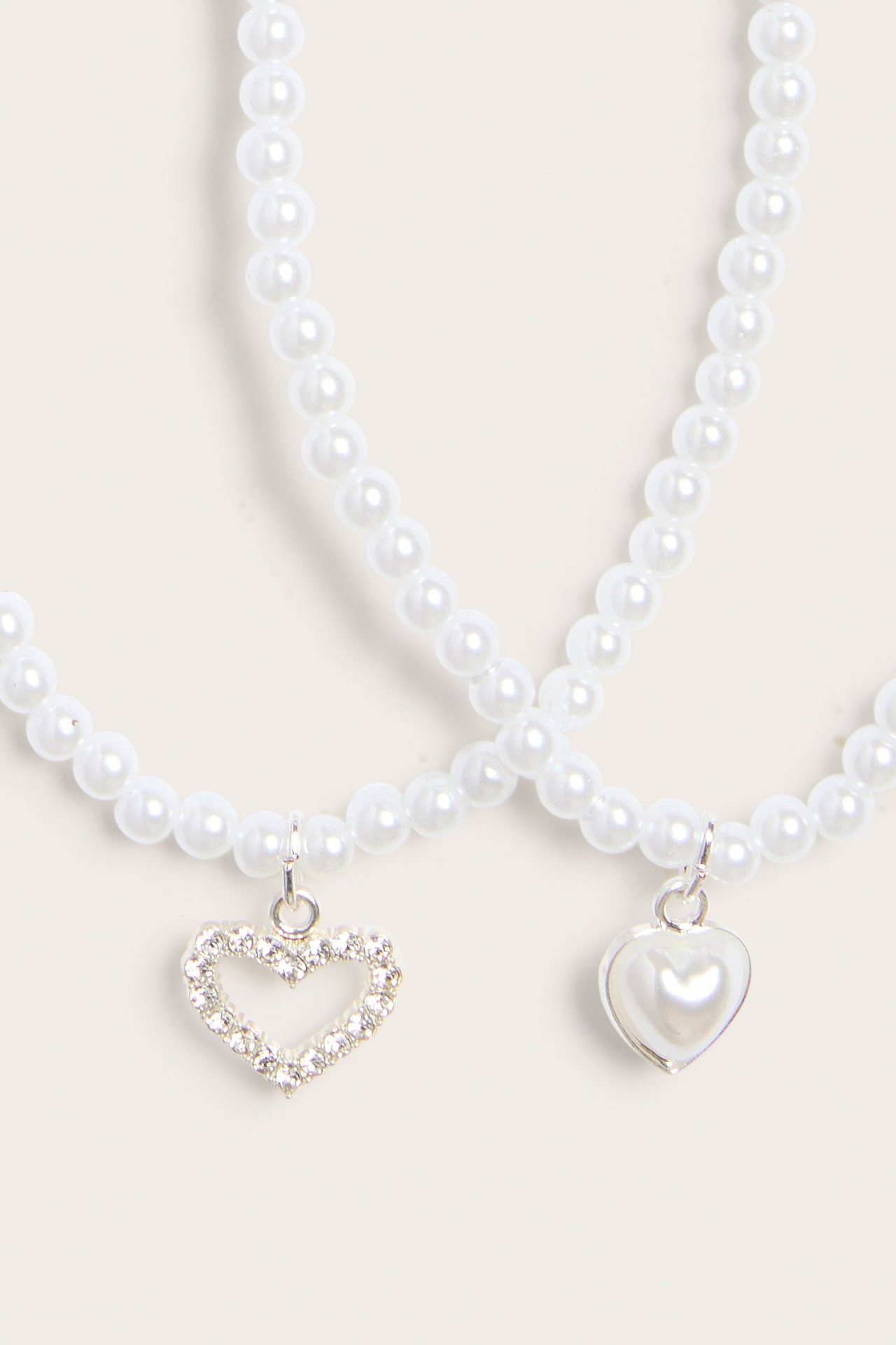 Lähikuva kahdesta luonnonvalkoisesta helmikaulakorusta, joissa sydänriipukset: toinen kimalteleva, toinen helmi.
