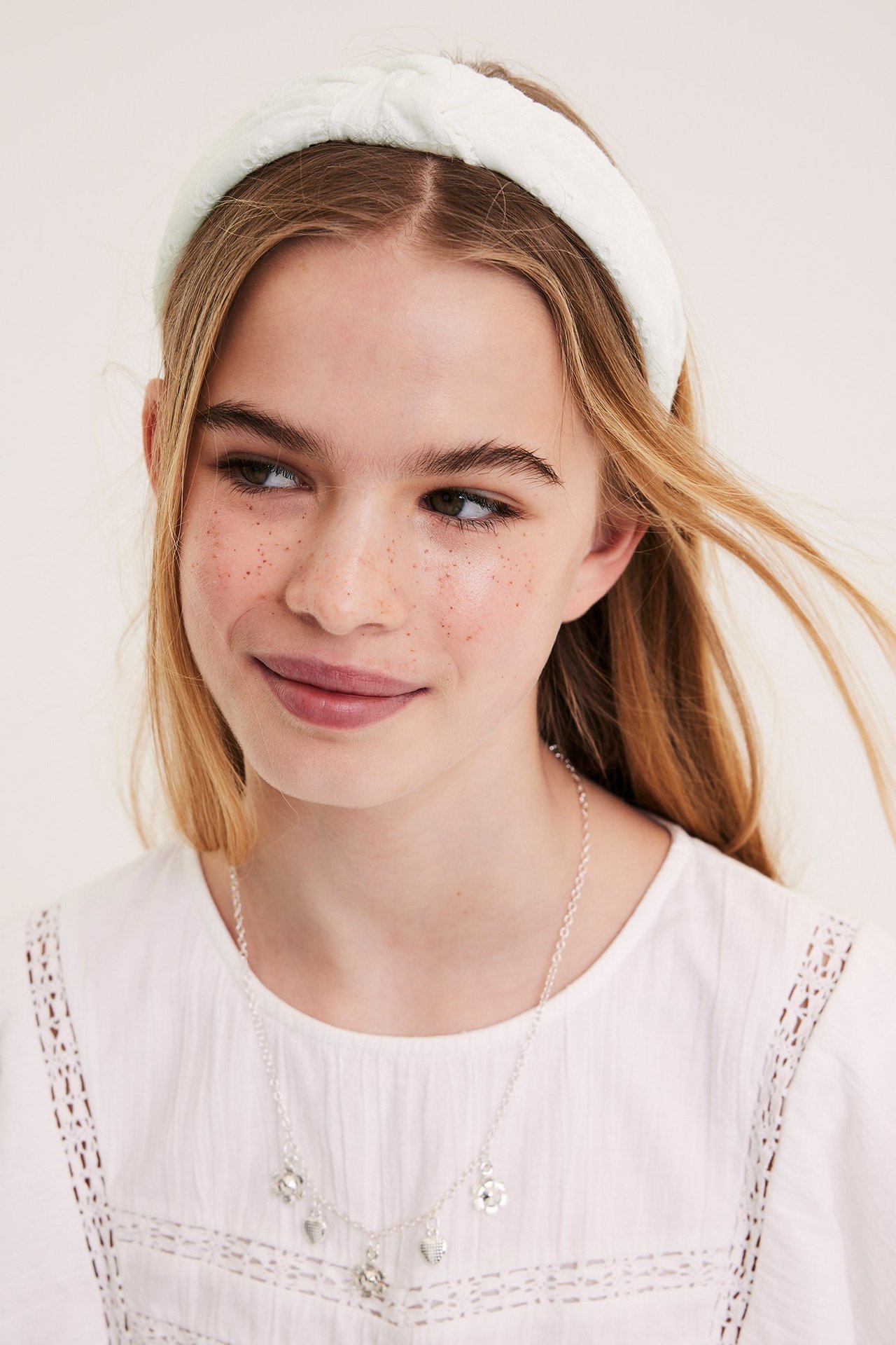 Edestä, lapsi pitää offwhite-kankaista broderie anglaise -pantaa, hopeista riipuskaulakorua ja valkoista toppia.