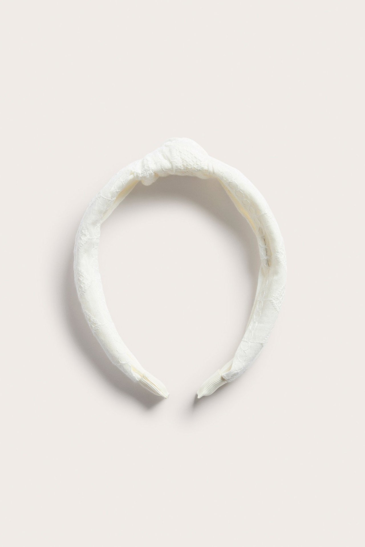 Framifrån: Ett offwhite tygdiadem med broderie anglaise och en knutdetalj.