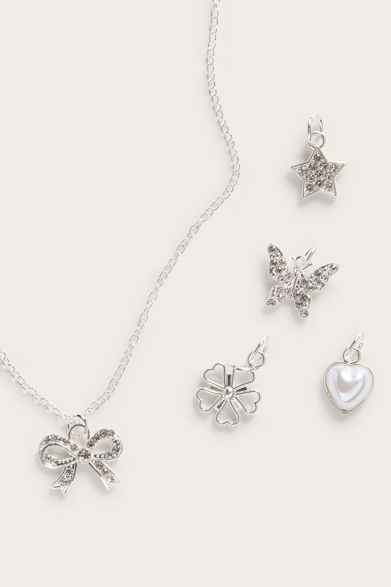 Närbild av silverfärgat halsband med strassrosettberlock, plus utbytbara berlocker: stjärna, fjäril, fyrklöver, pärlvit hjärta.