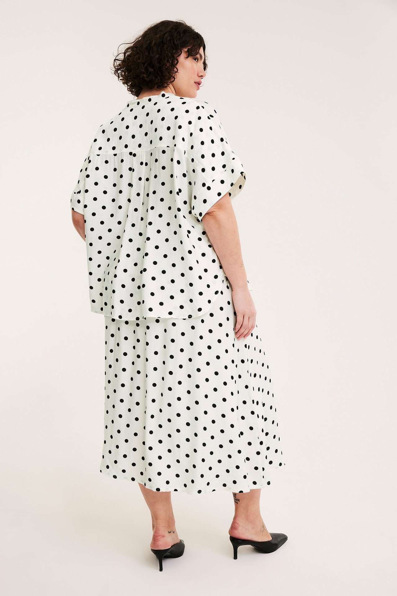 Kobieta, widok z tyłu, w kremowej spódnicy w kropki, pasującym luźnym topie i czarnych klapkach na obcasie.
