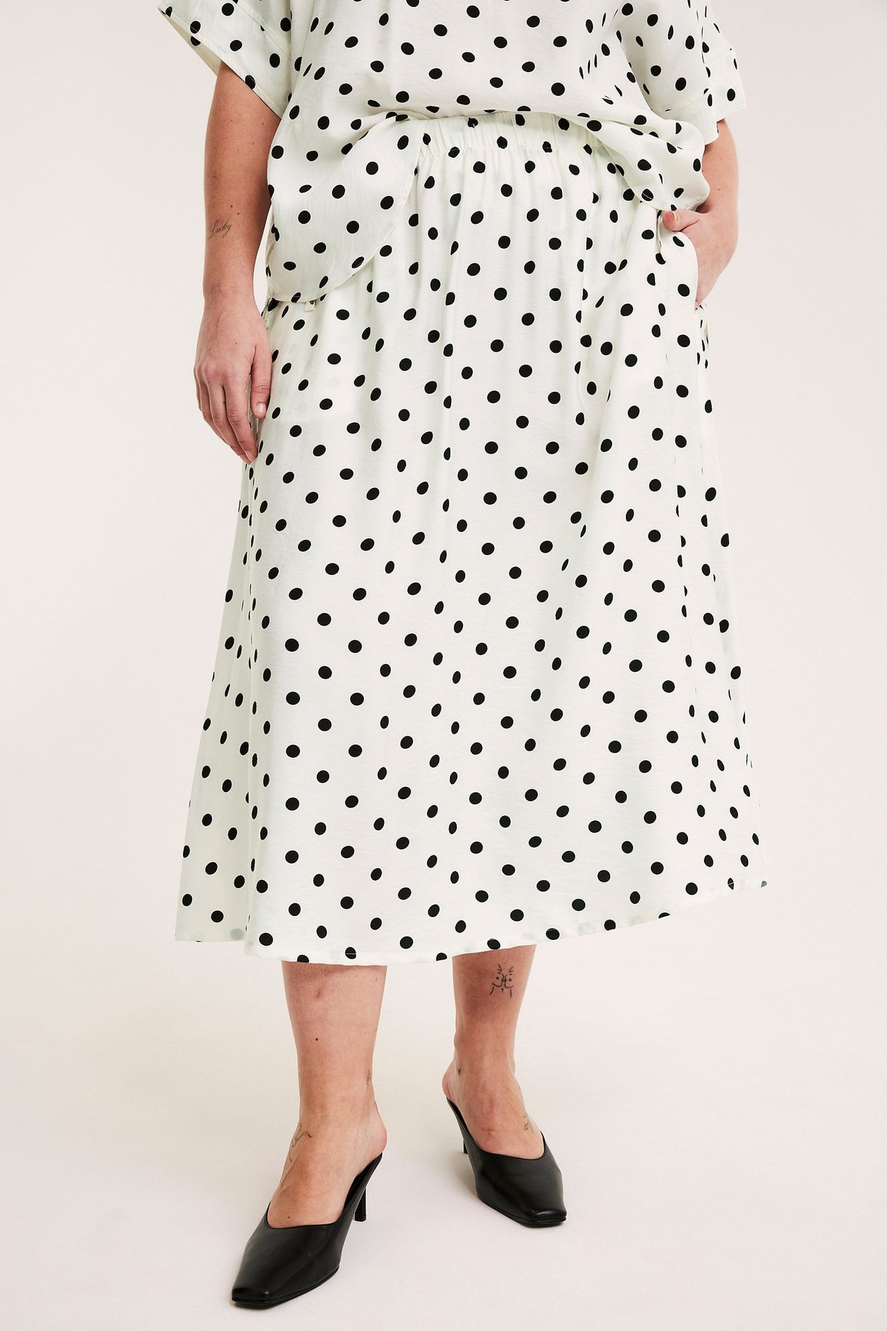 Frontbilde av en kvinne i et kremhvitt prikkete poplinskjørt, matchende topp og svarte mules.