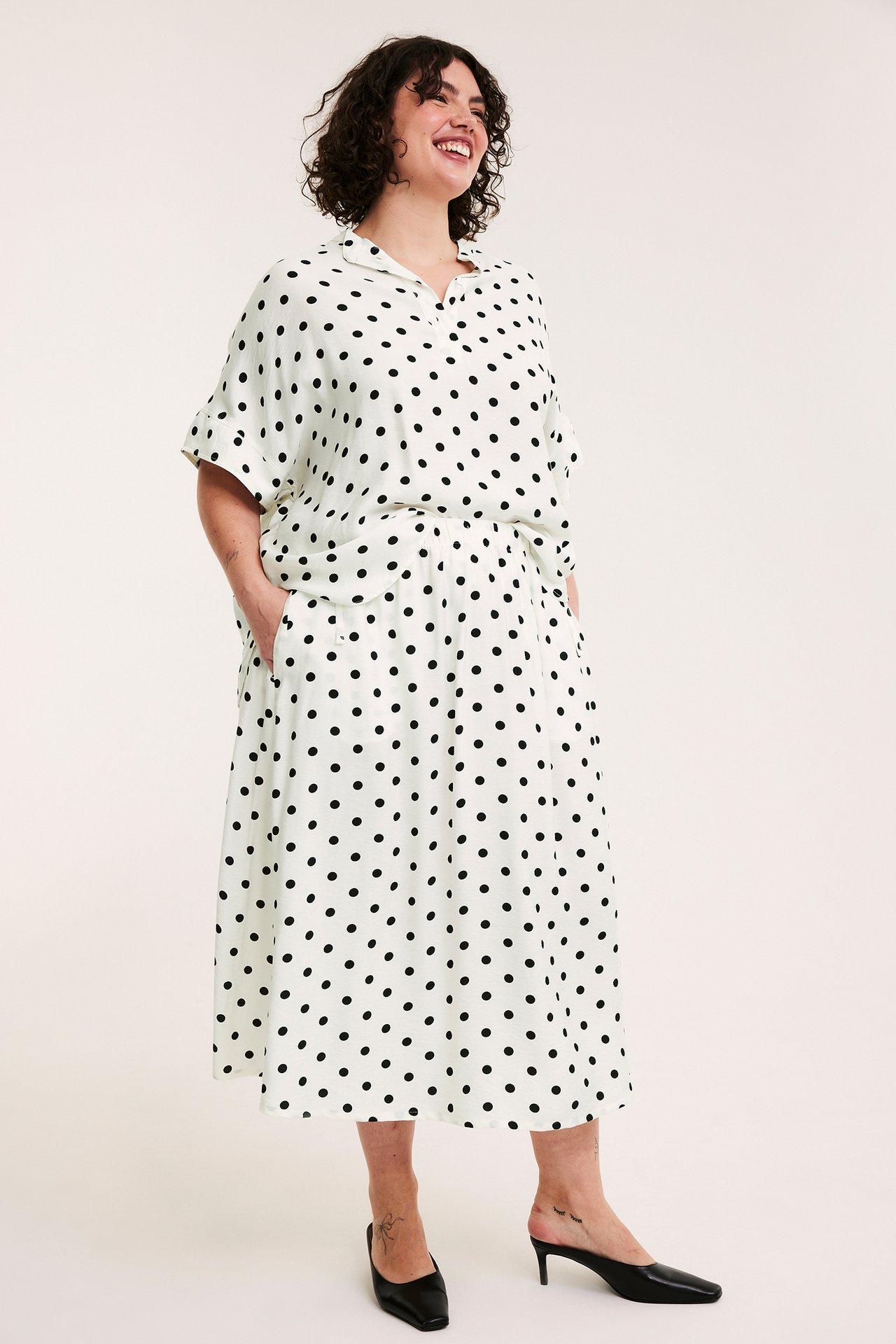 Forfra: Kvinne i et offwhite langt prikkete skjørt med elastisk midje og matchende bluse.