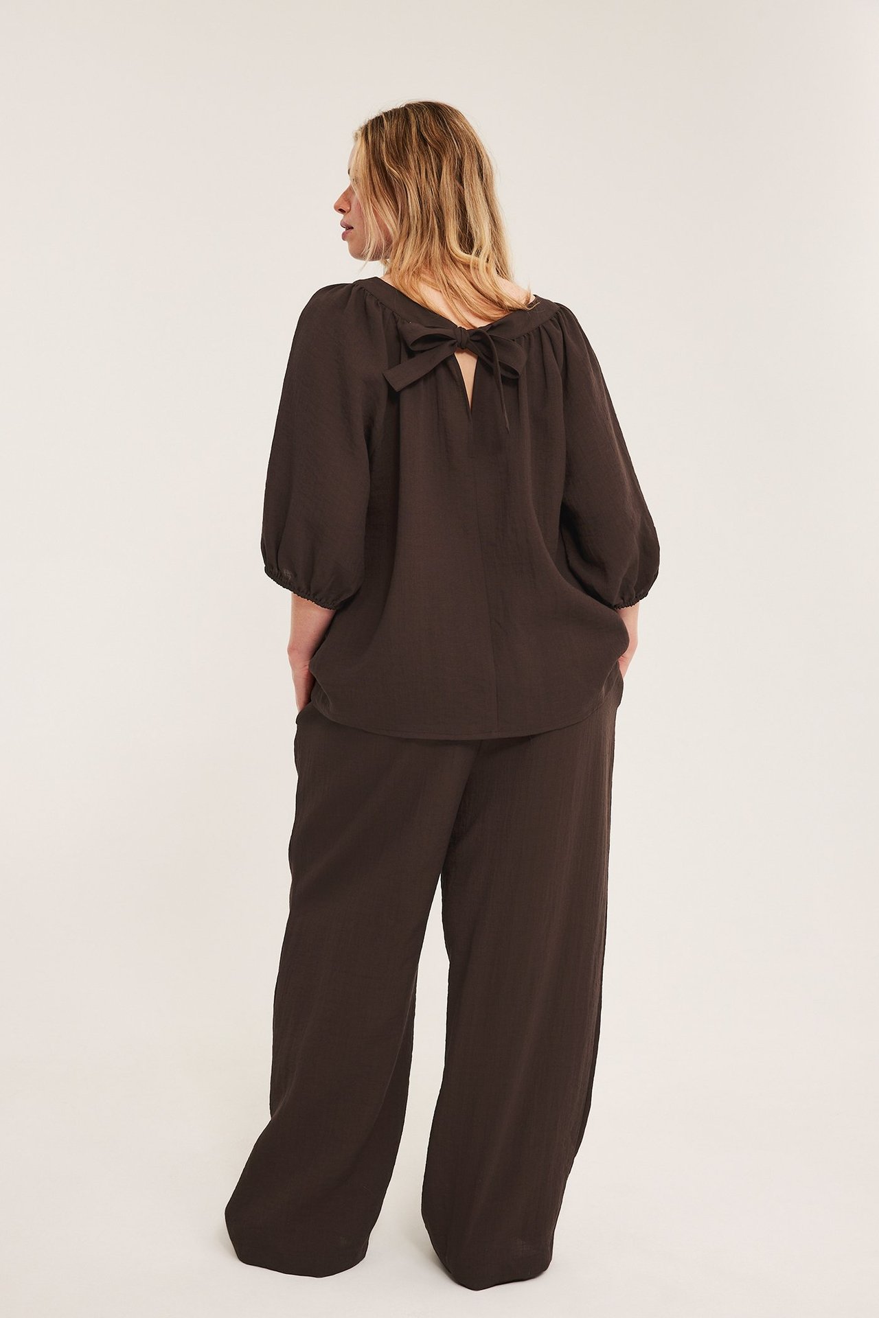 Bakfra, kvinne i mørkebrune vide bukser i viskoseblanding og matchende bluse med sløyfe bak.