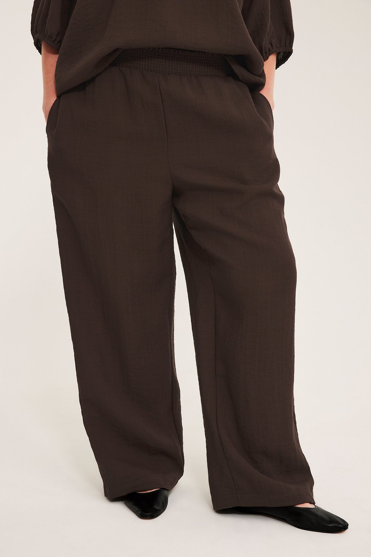 Edestä: Nainen yllään tummanruskeat leveät viskoosihousut smockvyötäröllä, taskuilla ja mustilla kengillä.