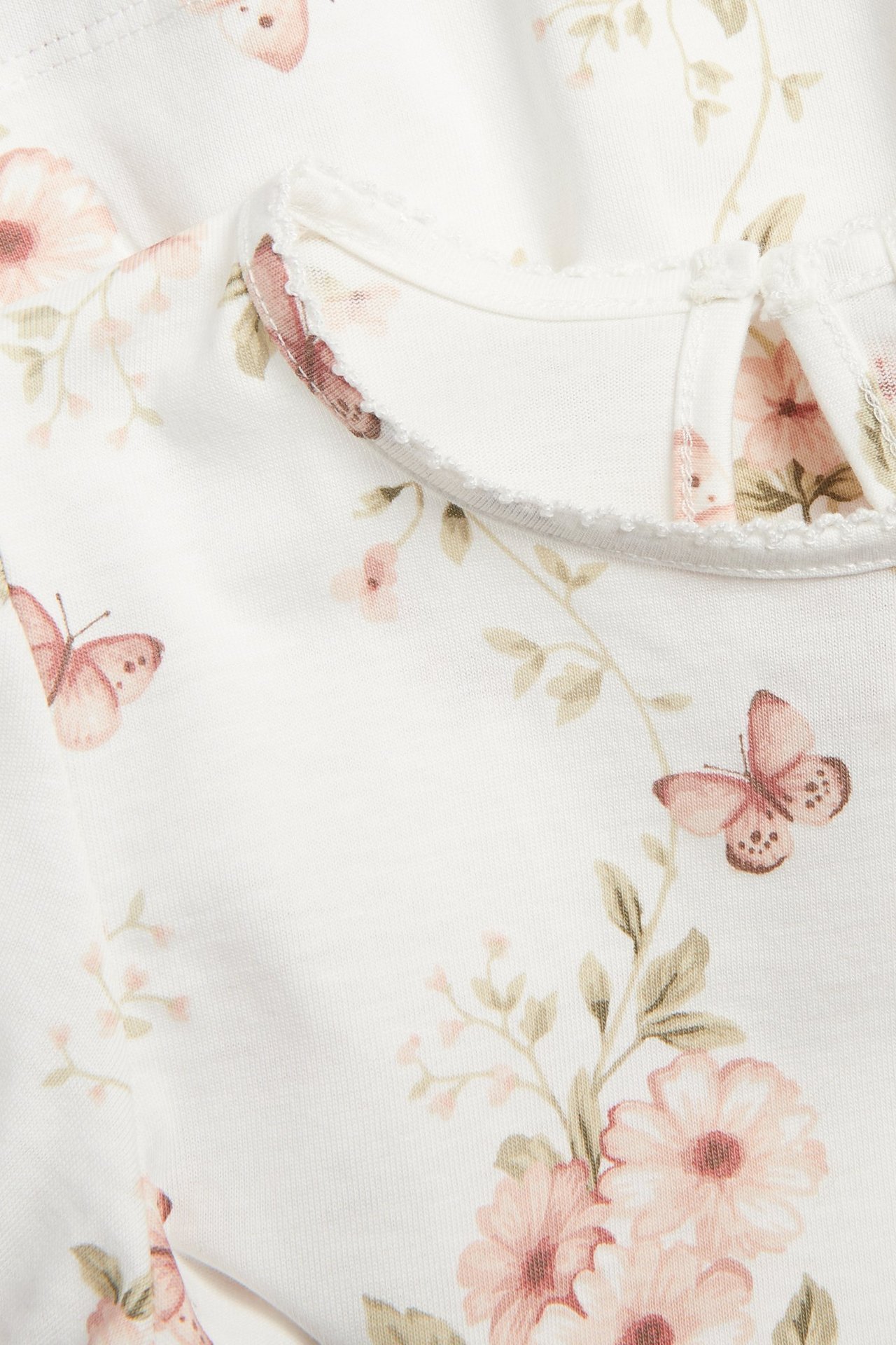Zbliżenie: biały bawełniany top z różowym kwiatowo-motylkowym wzorem, dekolt z pikotem, zapinany na guzik z tyłu.