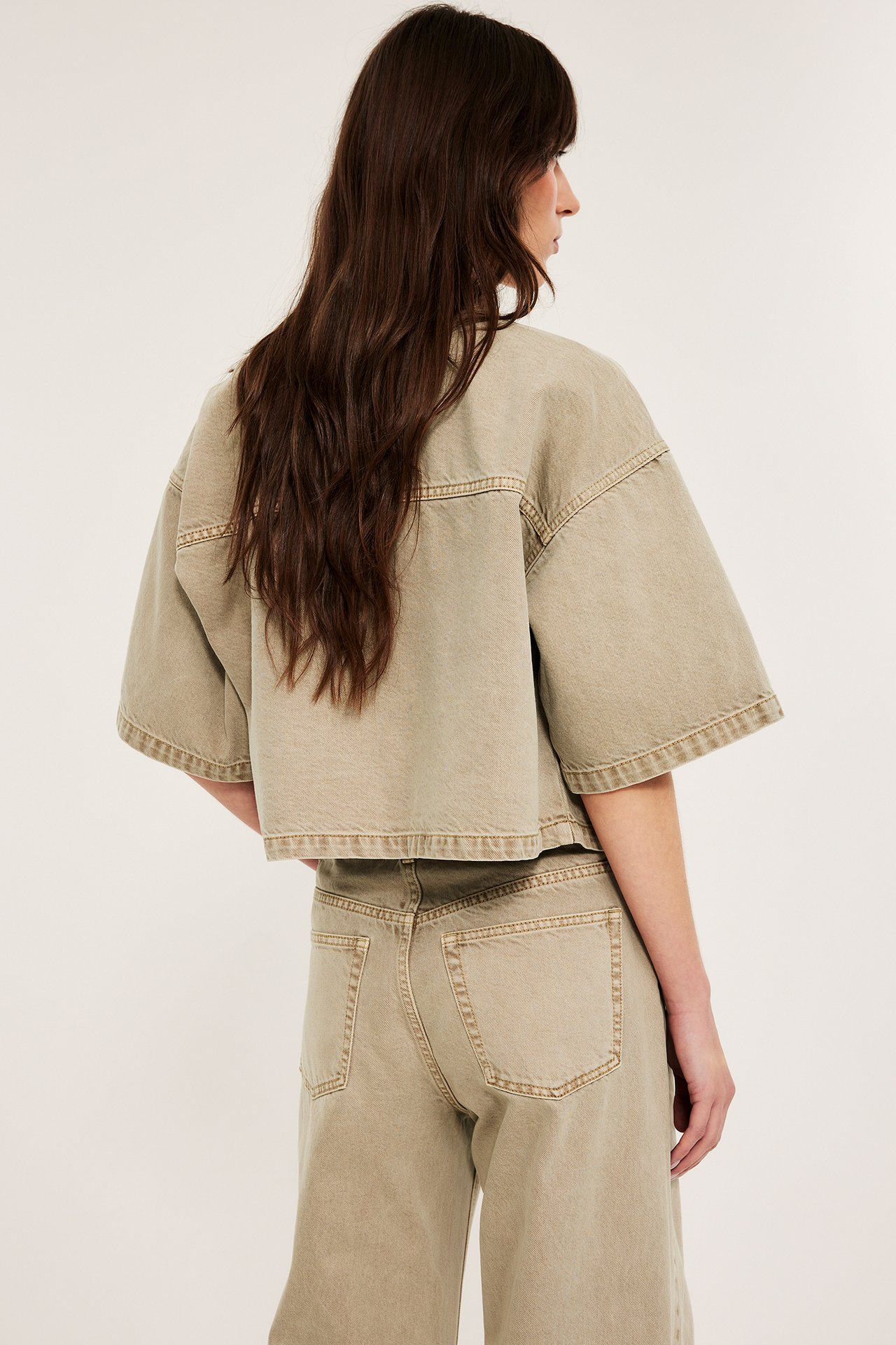 Widok z tyłu z boku: kobieta w jasnobrązowej krótkiej koszuli twillowej i pasujących szerokich jeansach.