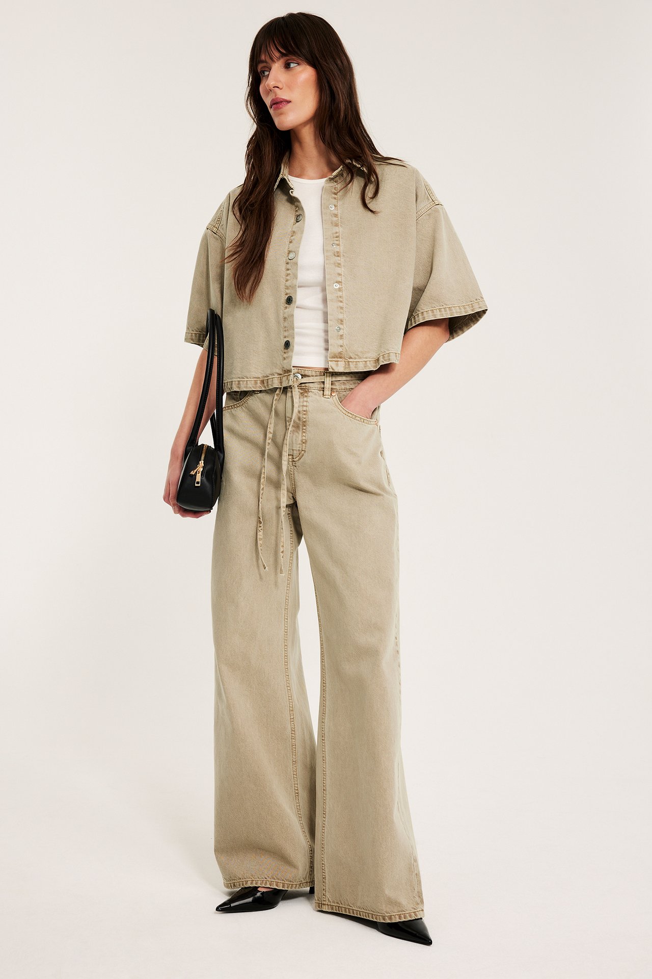 Frontbilde: Kvinne i lysbrun kort twillskjorte, vide bukser, hvit topp, svarte sko, holder håndveske.