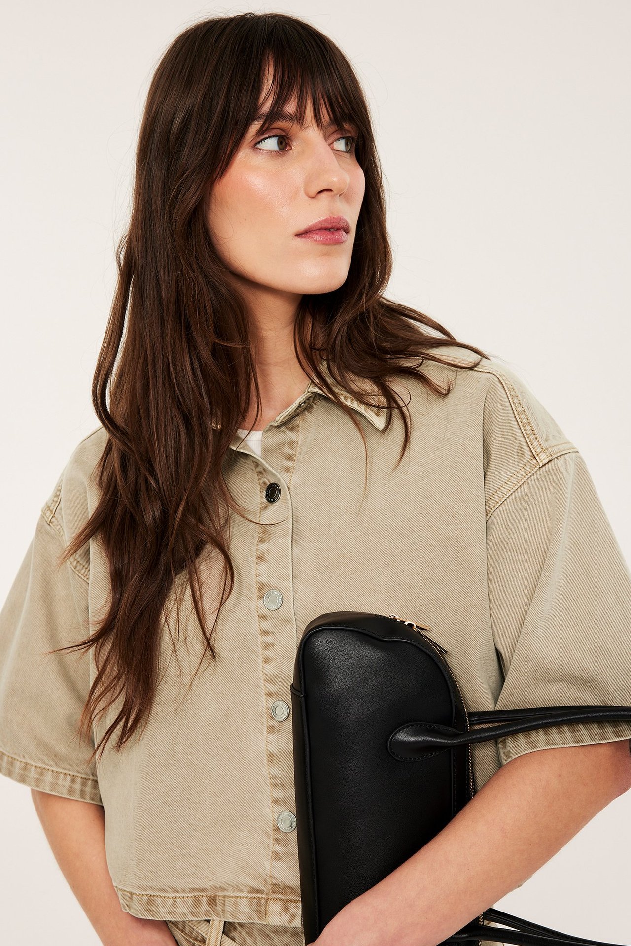 Närbild av kvinna i ljusbrun kortärmad twillskjorta med jeansknappar, hållande en svart handväska.