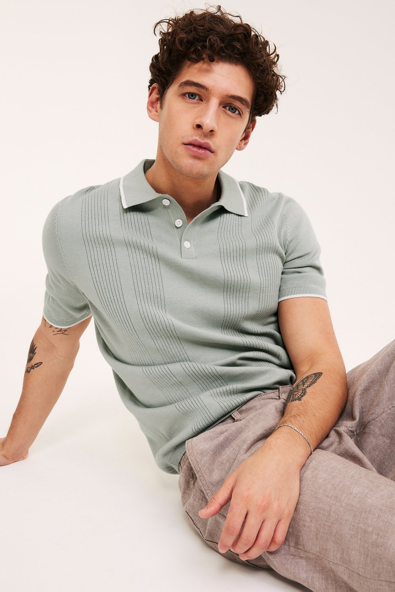 Framifrån, man i ljusgrön stickad pikétröja med vertikala ränder, vit kant, och bruna byxor.