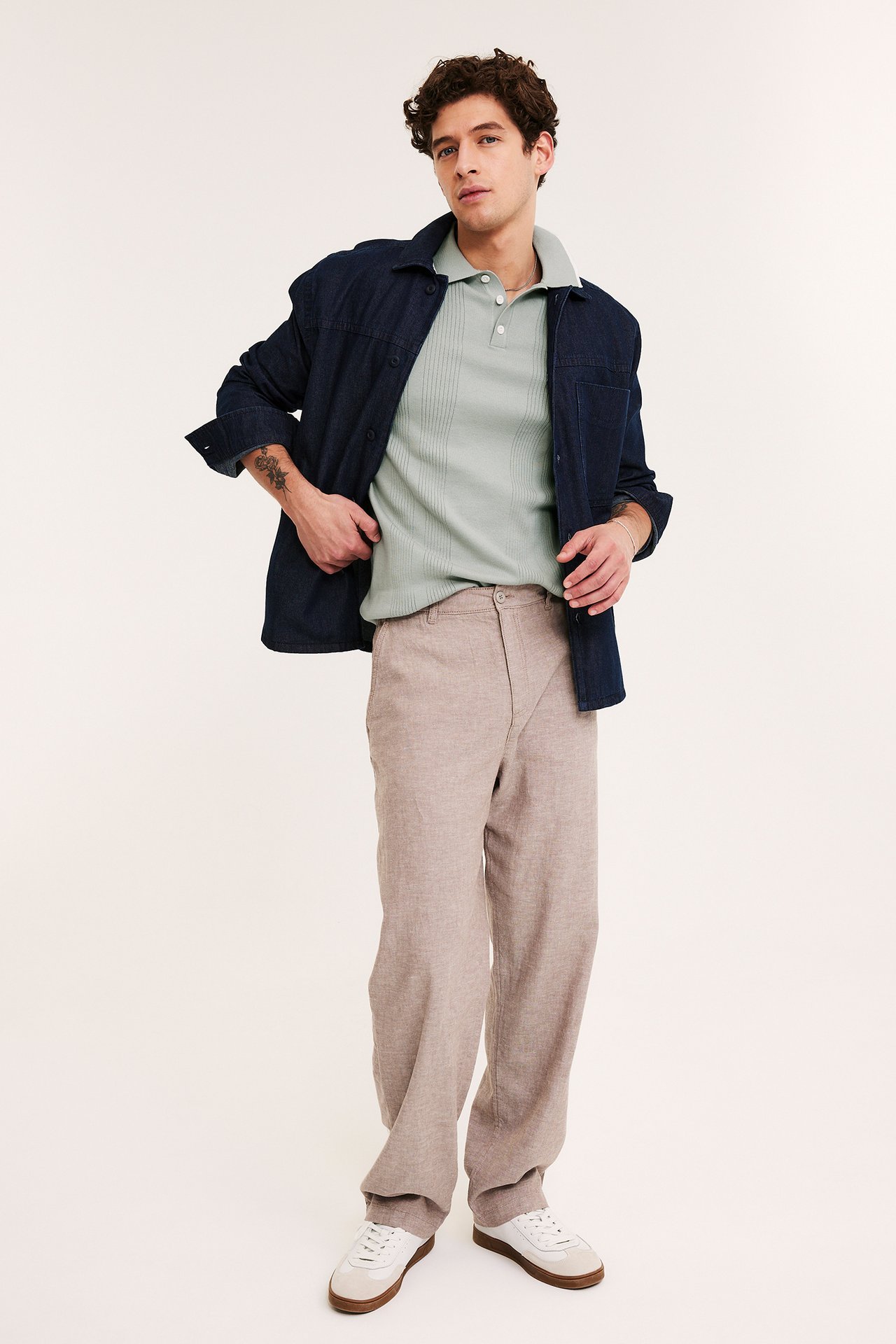 Edestä: Mies vihreässä ribbineulepoolopaidassa, tummansinisessä farkkupaidassa, beigeissä housuissa ja valkoisissa tennareissa.