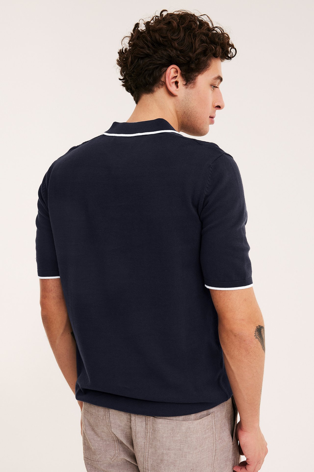 Widok z tyłu: Mężczyzna w granatowej dzianinowej koszulce polo z białym wykończeniem kołnierza i mankietów, oraz jasnobrązowych szortach.