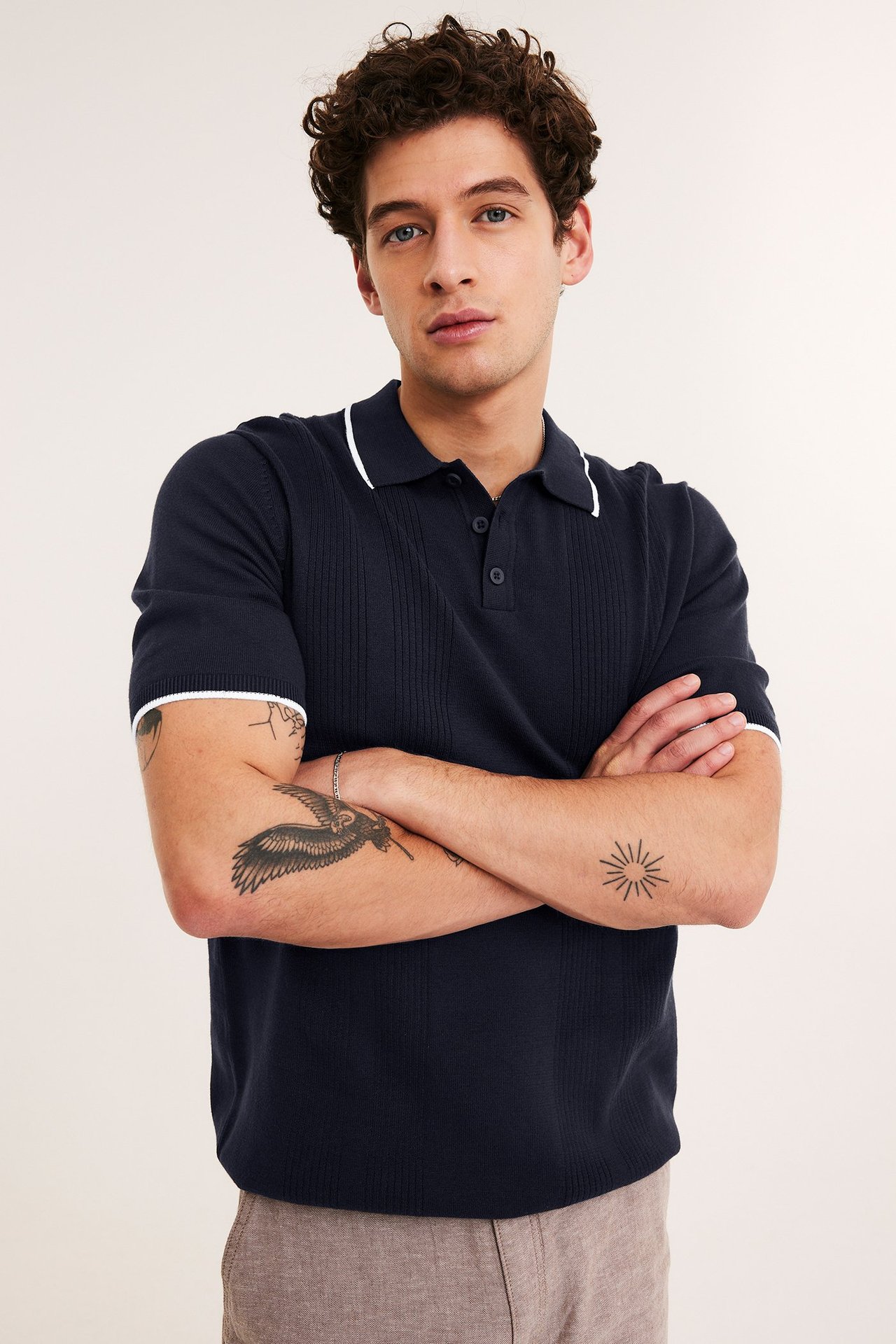 Mann, forfra, i en mørkeblå strikket pologenser med hvit kant og lysebrune bukser.