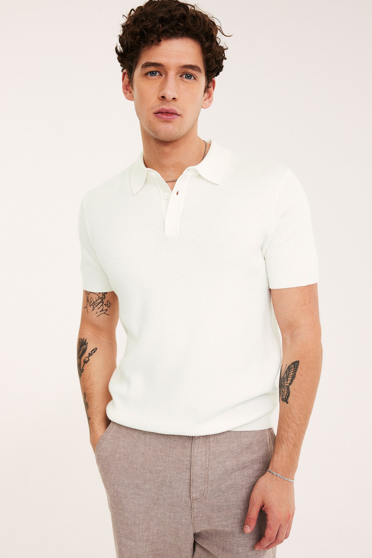 Edestäpäin: mies yllään luonnonvalkoinen struktuurineulottu pikeepaita kolmella napilla, ruskeat housut ja hopearannekoru.