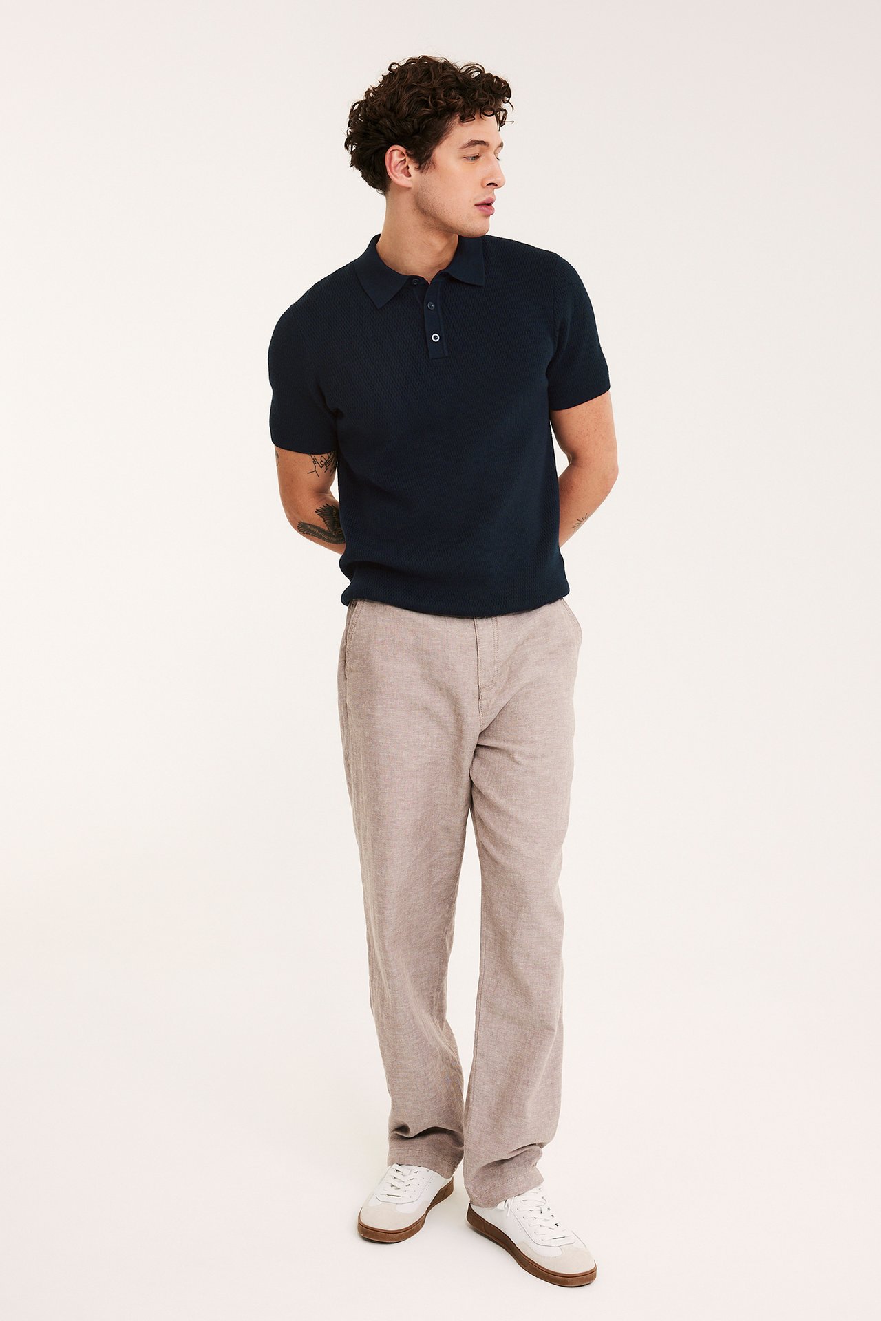 Edestä päin kokokuva miehestä tummansinisessä neulotussa pikeepaidassa, vaaleanruskeissa housuissa ja valkoisissa lenkkareissa.