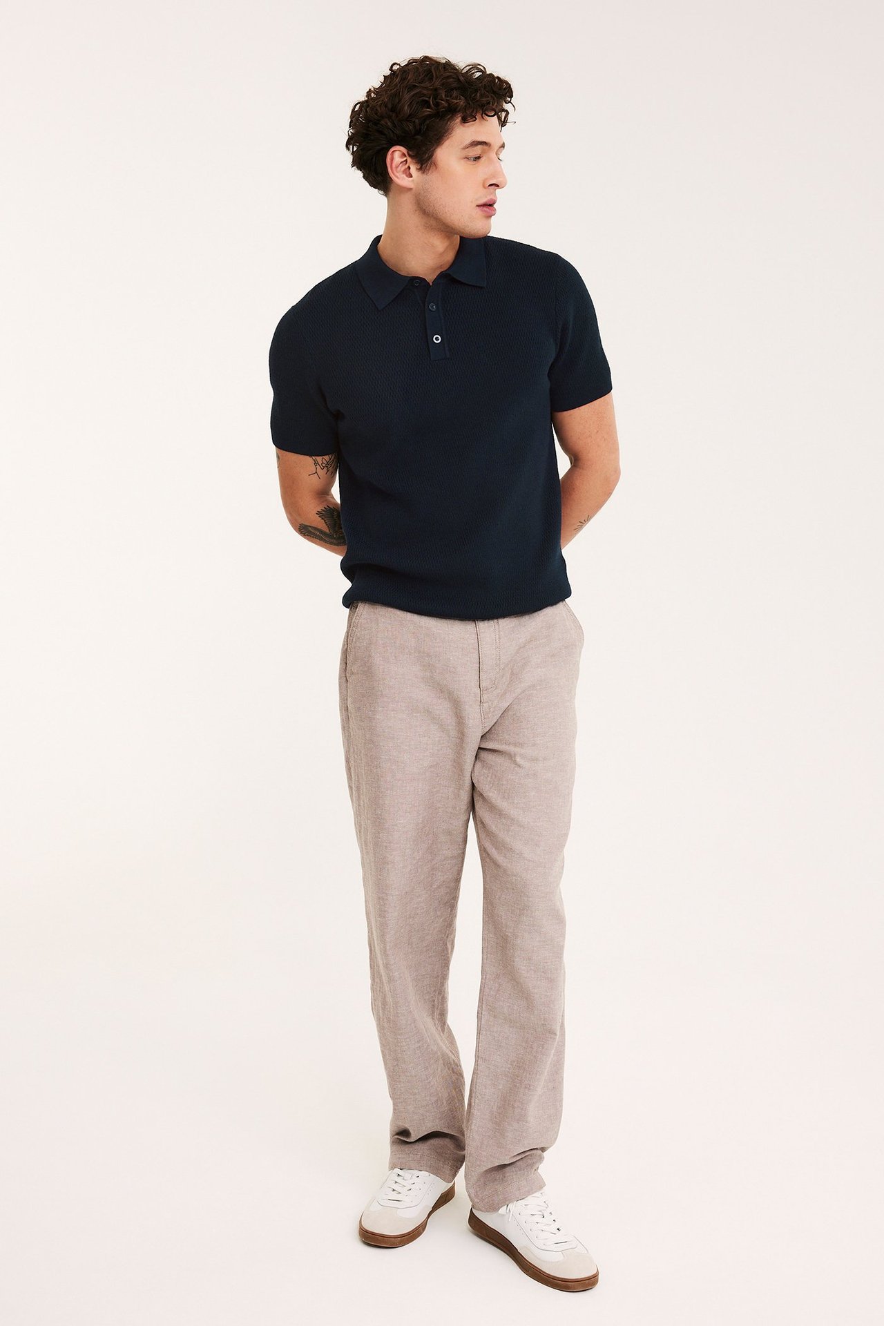 Helkroppsvy framifrån av en man i mörkblå stickad pikétröja, ljusbruna byxor och vita sneakers.