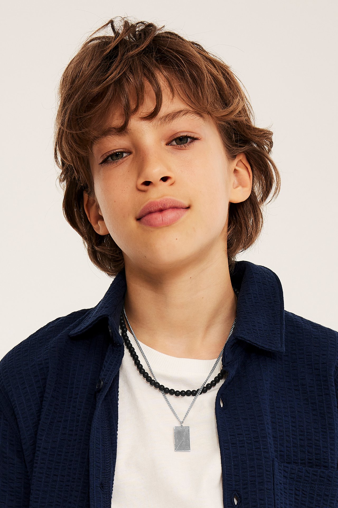 Zbliżenie dziecka w białym T-shircie i granatowej koszuli, noszącego podwójny srebrny naszyjnik z wisiorkiem i czarnymi koralikami.