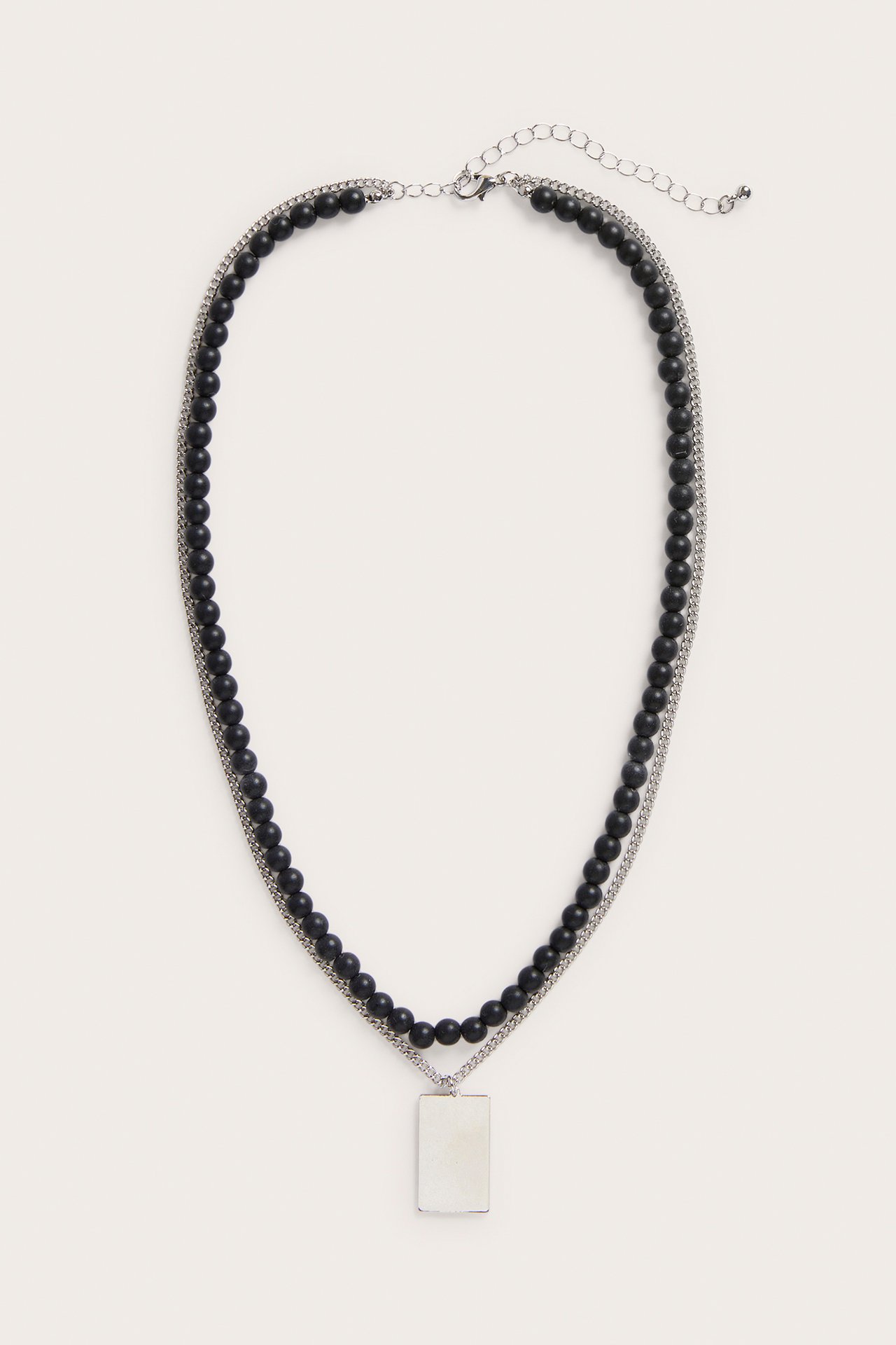 Frontbilde av et sølvfarget dobbelt kjede med svarte perler og et rektangulært sølvfarget anheng, med karabinlås.