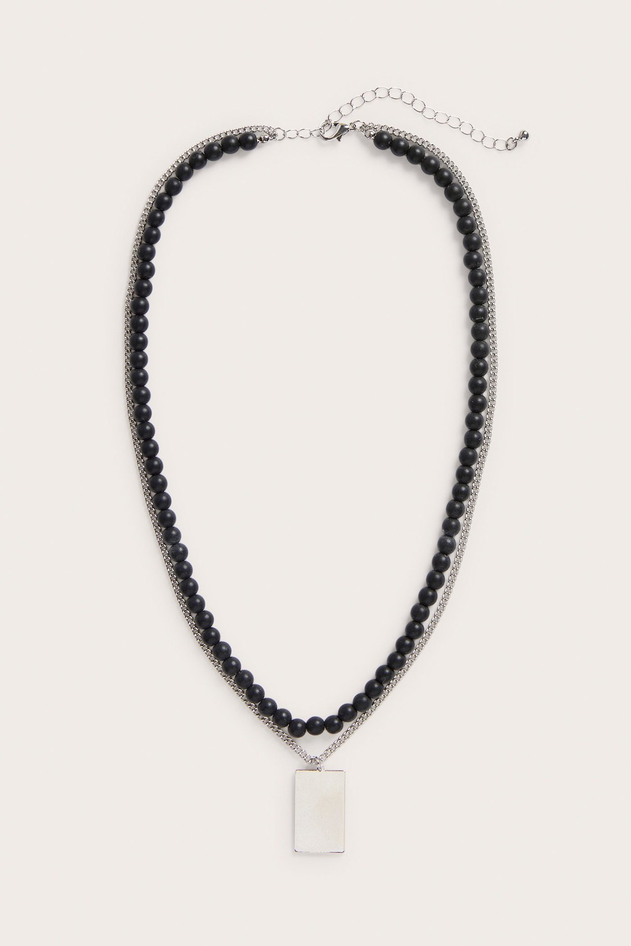 Widok z przodu srebrnego podwójnego naszyjnika z czarnymi koralikami i prostokątną srebrną zawieszką, z zapięciem karabinkowym.