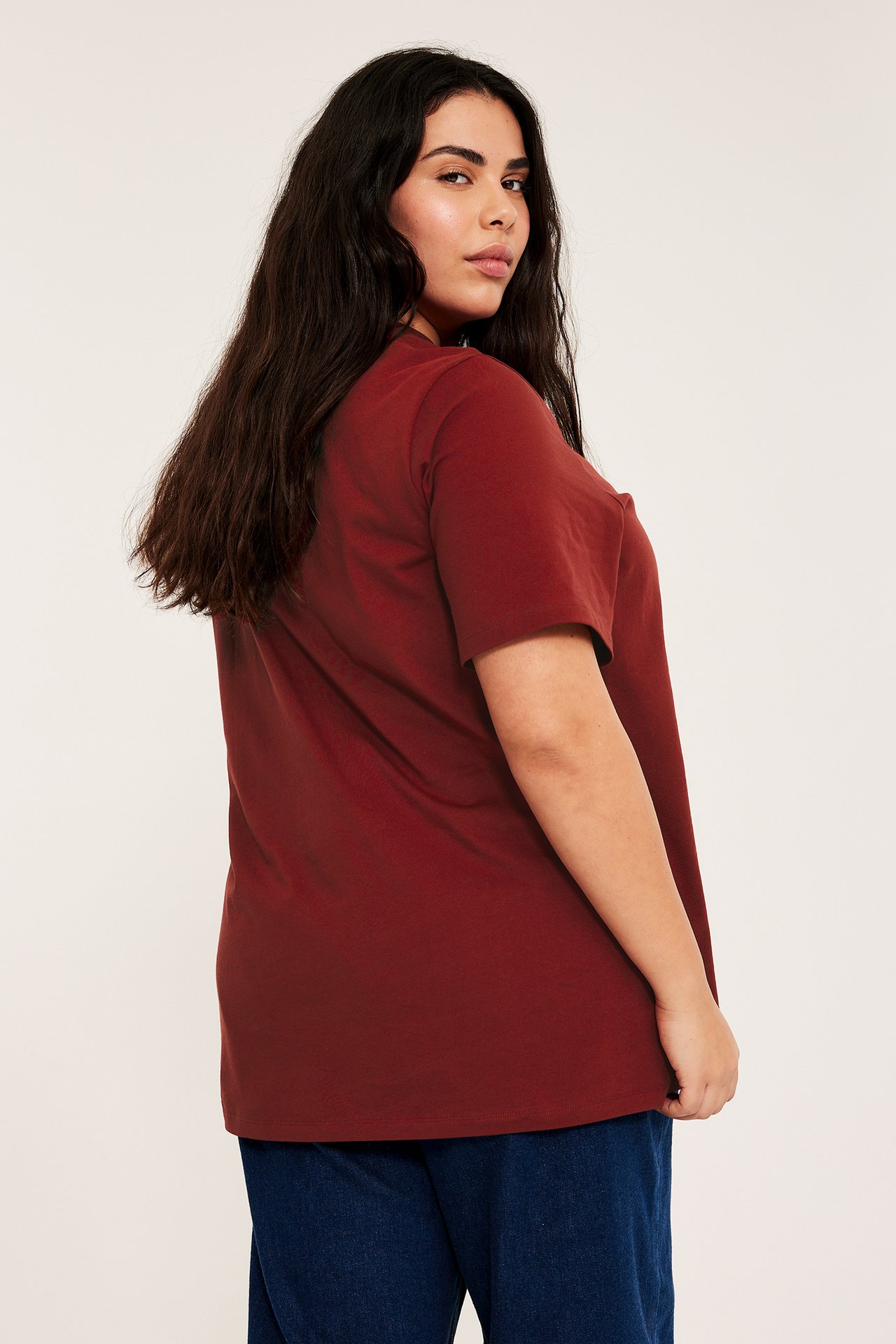 Bakside av en kvinne i en ensfarget mørkerød lang bomull-t-skjorte og blå jeans.