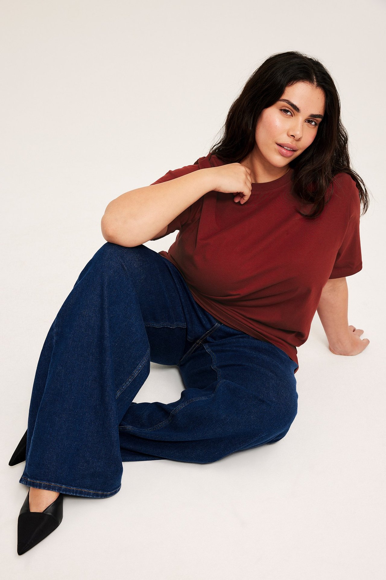 Forfra, kvinne i løs rød bomullslang t-skjorte, blå vide jeans og svarte spisse flate sko.