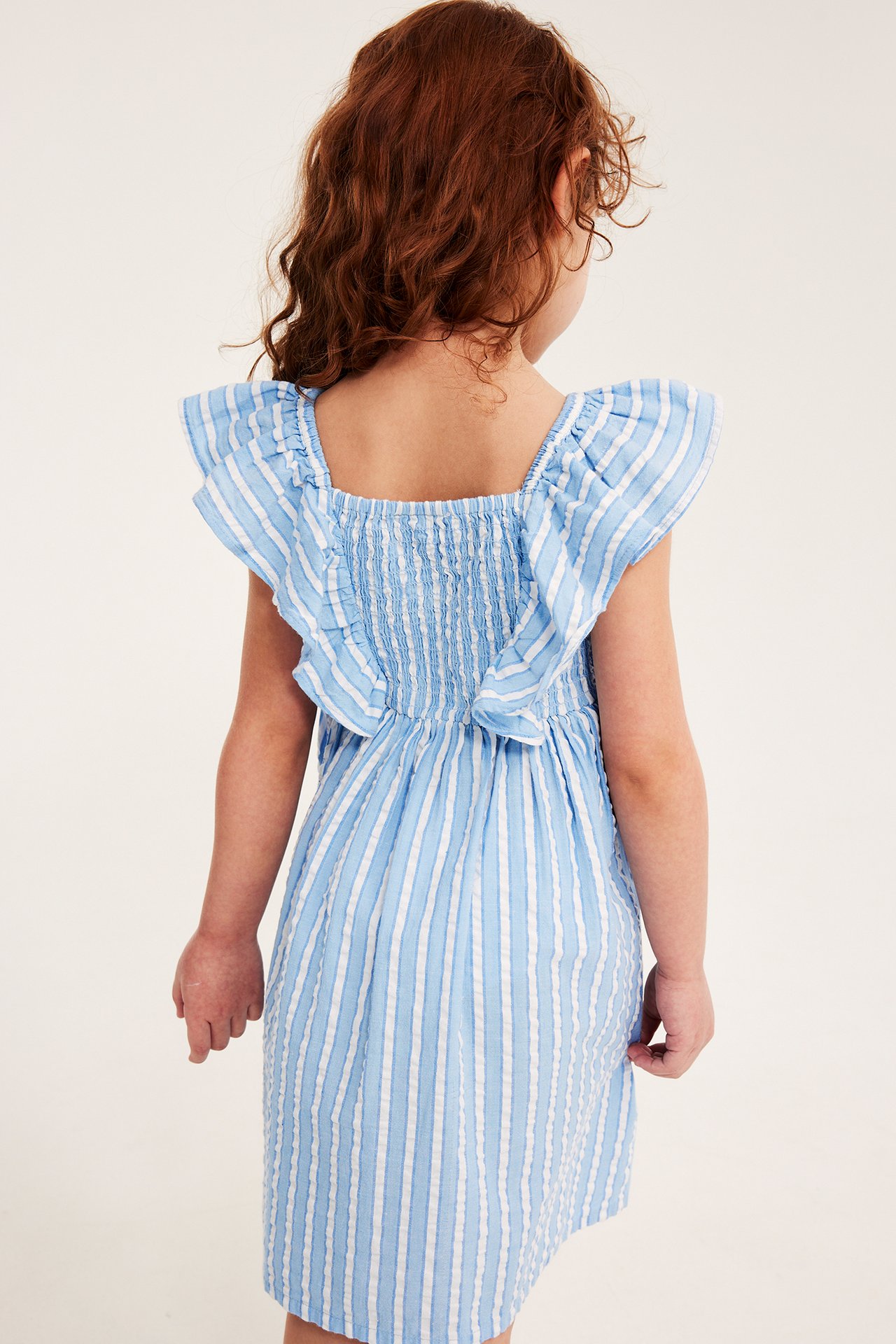 Widok z tyłu: dziecko w jasnoniebiesko-białej bawełnianej sukience w paski, z falbanami i marszczoną górą.