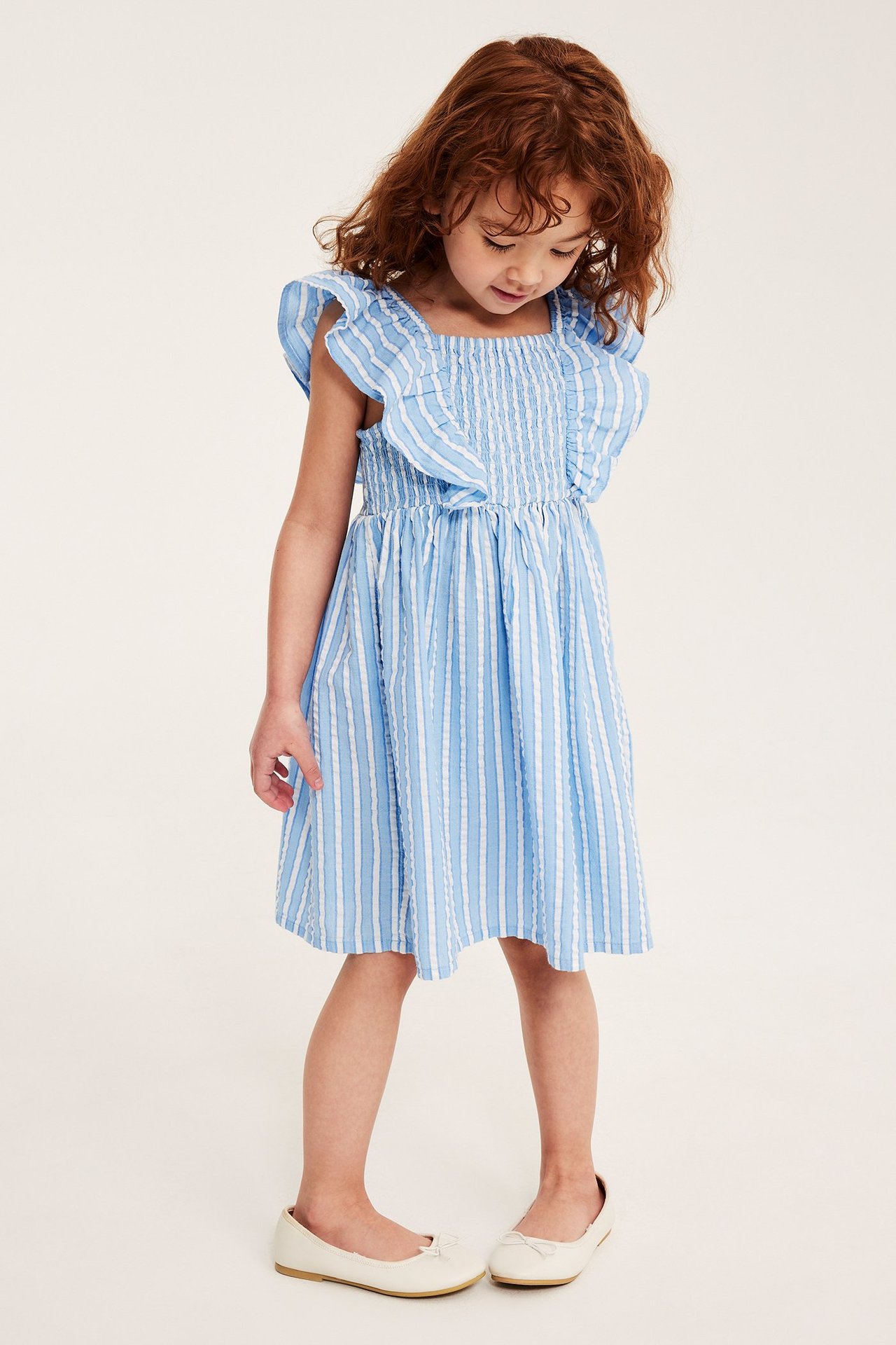 Edestäpäin näkymä lapsesta yllään vaaleansininen raidallinen seersucker-mekko röyhelöhihoilla ja vaaleanbeiget ballerinakengät.